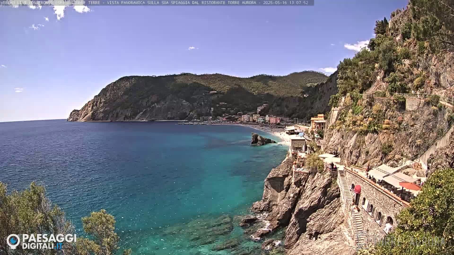 A sunny coastal view shows a town nestled in a cove, with a rocky cliff and staircase leading down to a sandy beach, and clear turquoise water.