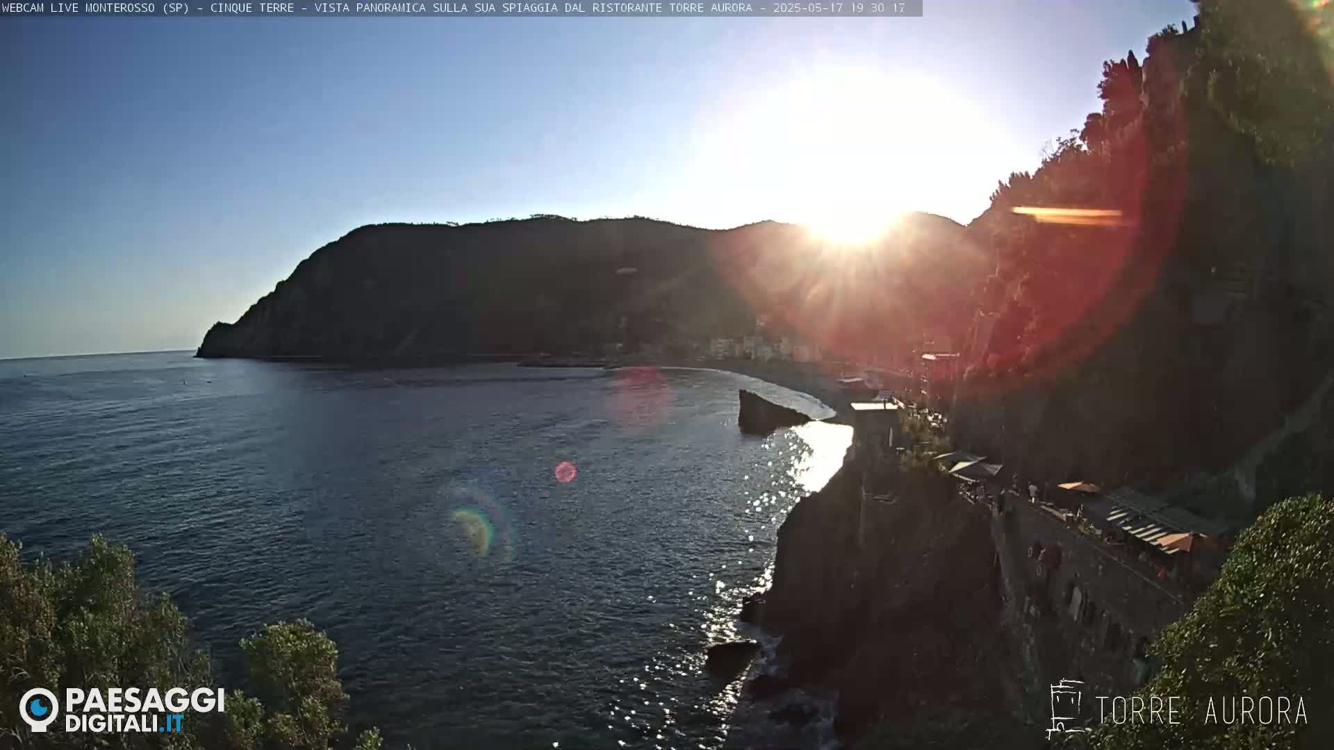 A coastal town nestled between a rocky cliff and the sea is bathed in the golden light of a setting sun.