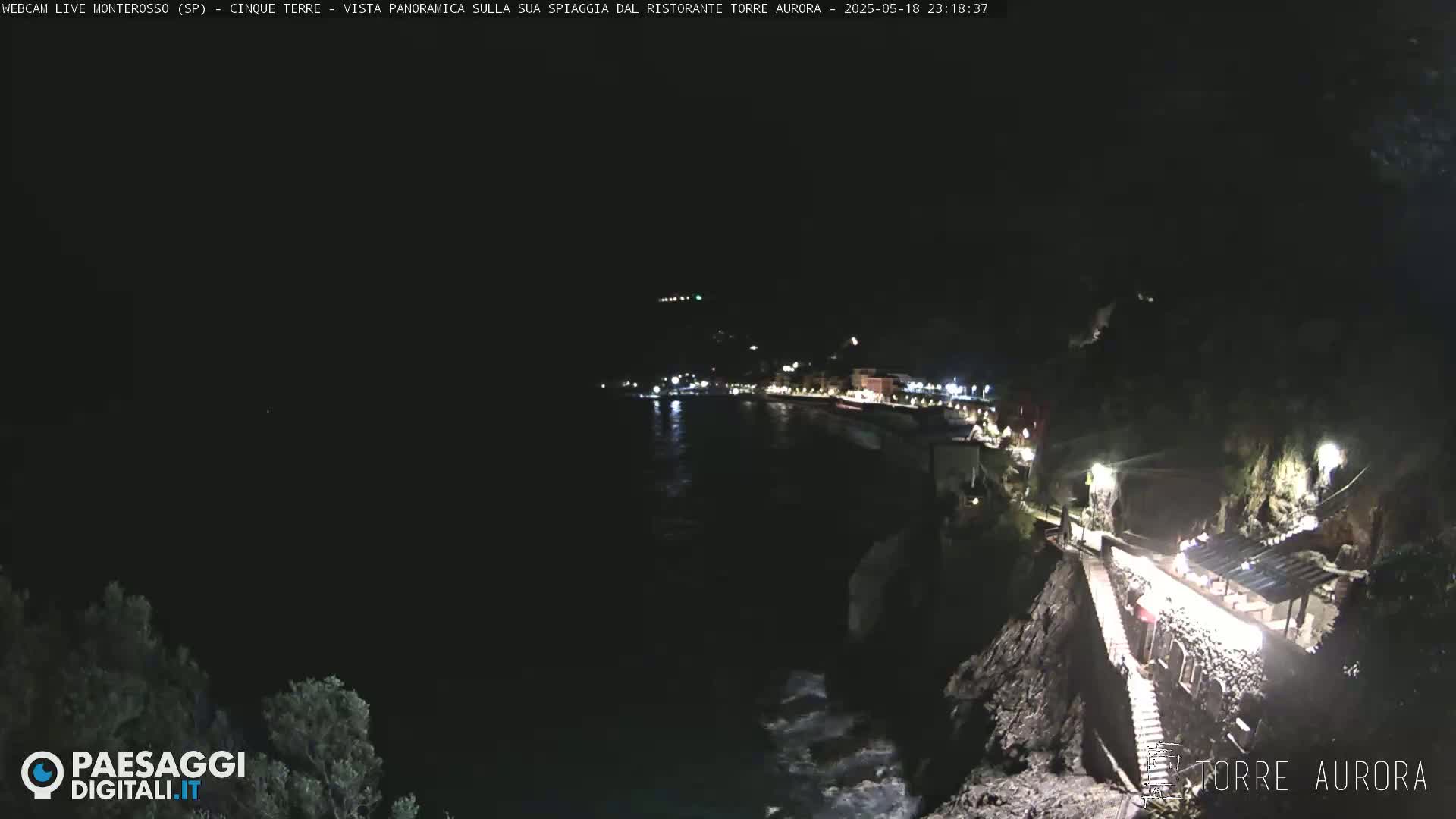 A nighttime coastal scene shows a dark ocean with small waves lapping against a rocky shore, where illuminated buildings and walkways are visible along the cliffside.