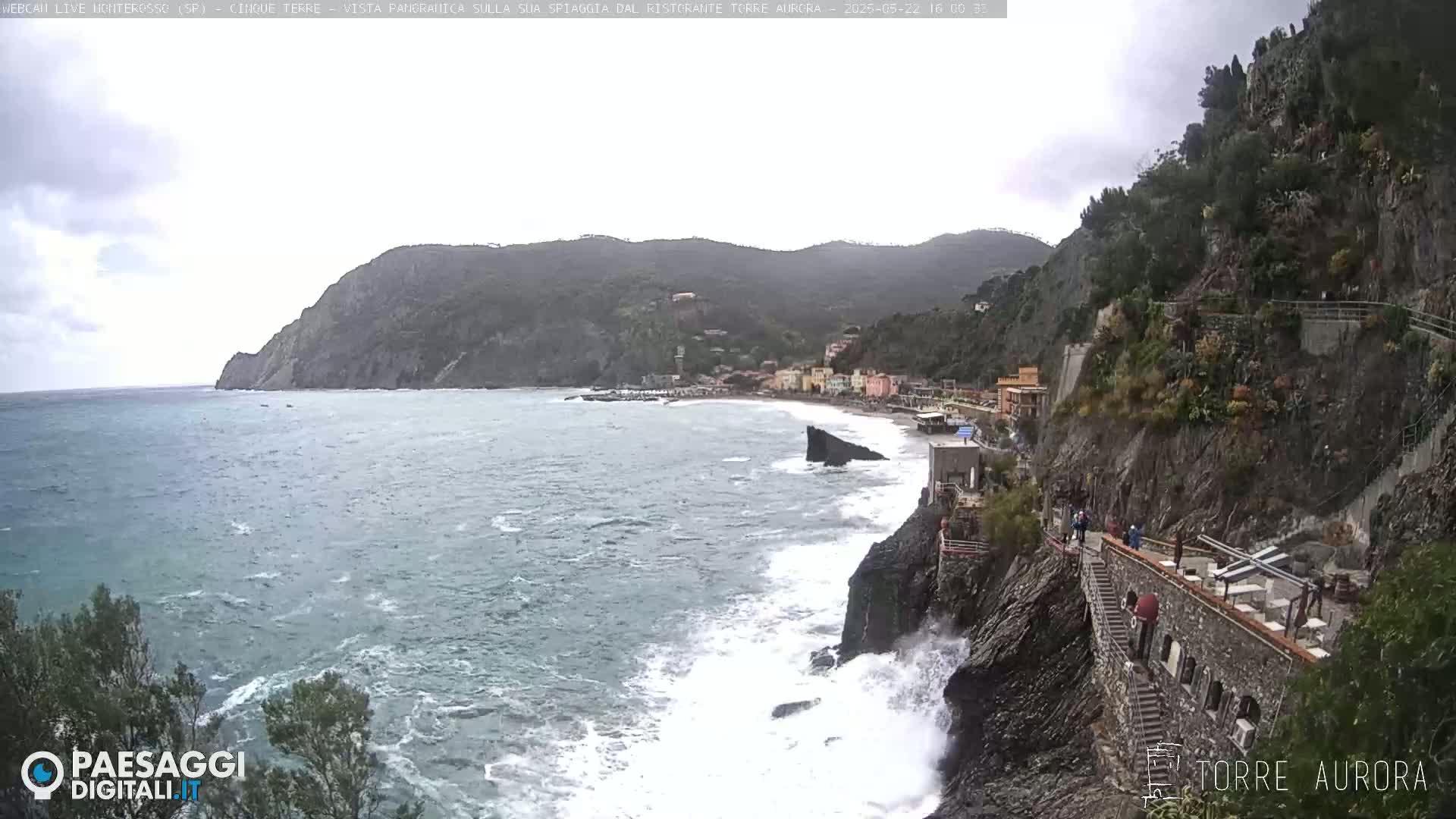 A coastal town nestled between a steep, green cliff and a rough, gray sea under an overcast sky.