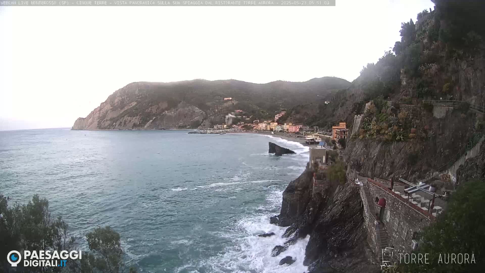 A coastal town nestled between steep cliffs overlooks a rocky beach with crashing waves under an overcast sky.