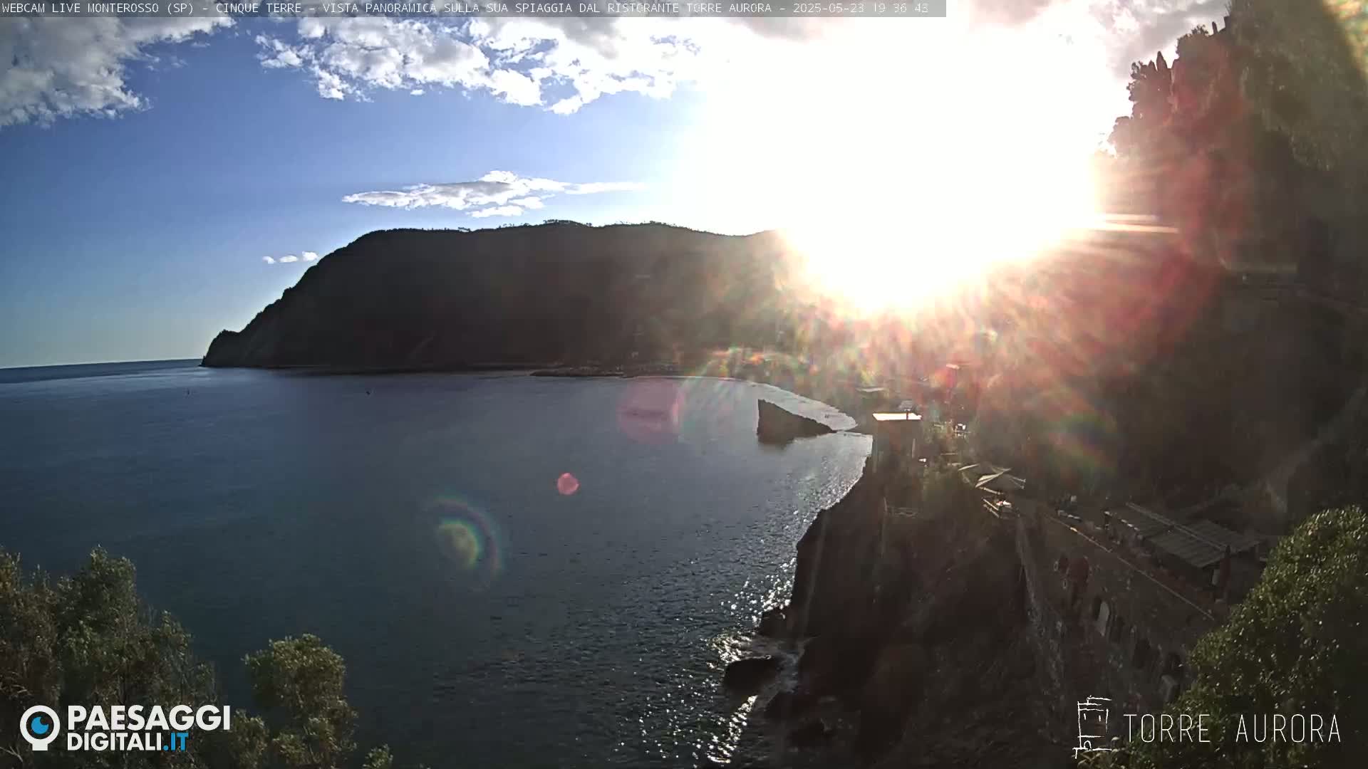 A sun-drenched coastal scene shows a calm bay with a rocky headland and distant hills under a partly cloudy sky.