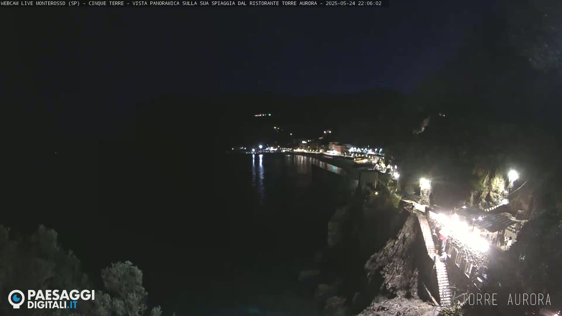 A nighttime, coastal view shows a dark body of water reflecting lights from a town on a hillside, with a lit pathway and structure descending from the town towards the water.