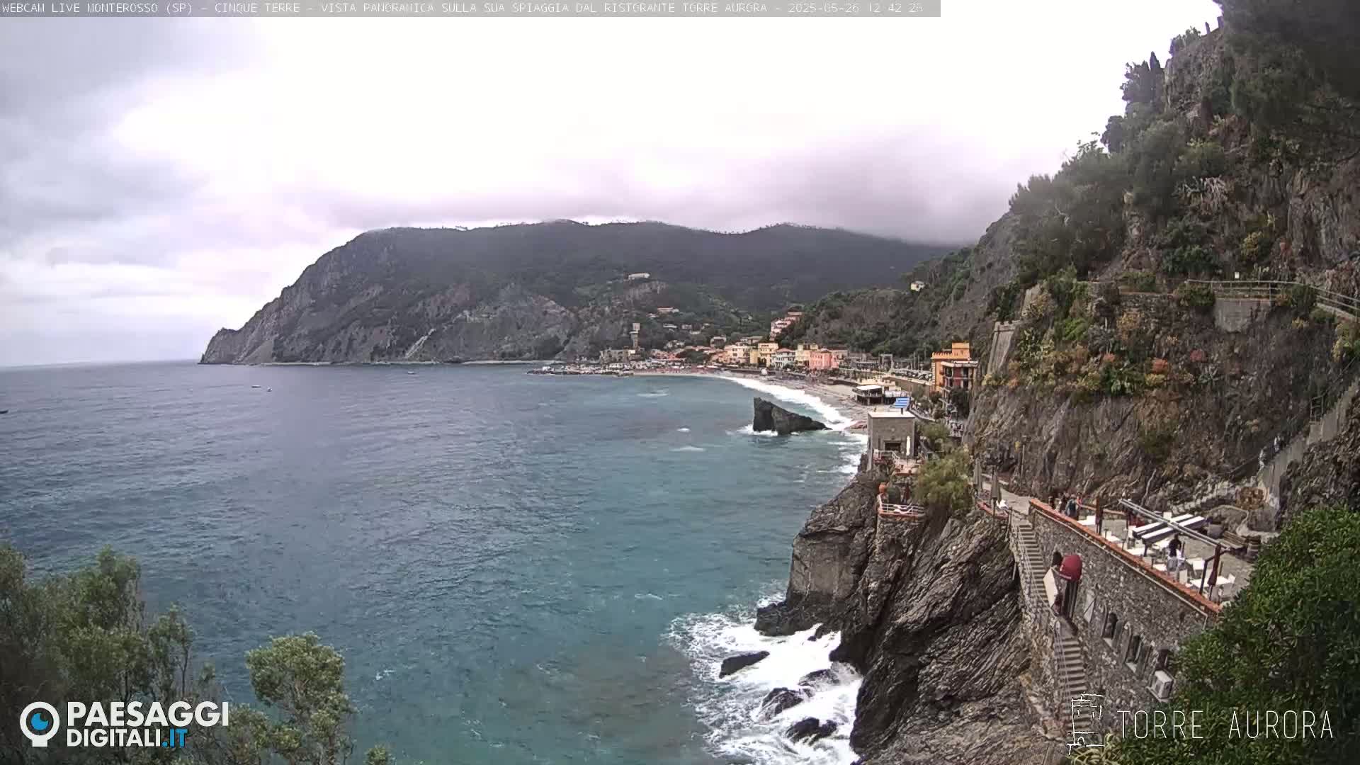 A coastal town nestled between a rocky cliff and the sea on an overcast day, with a beach visible below.