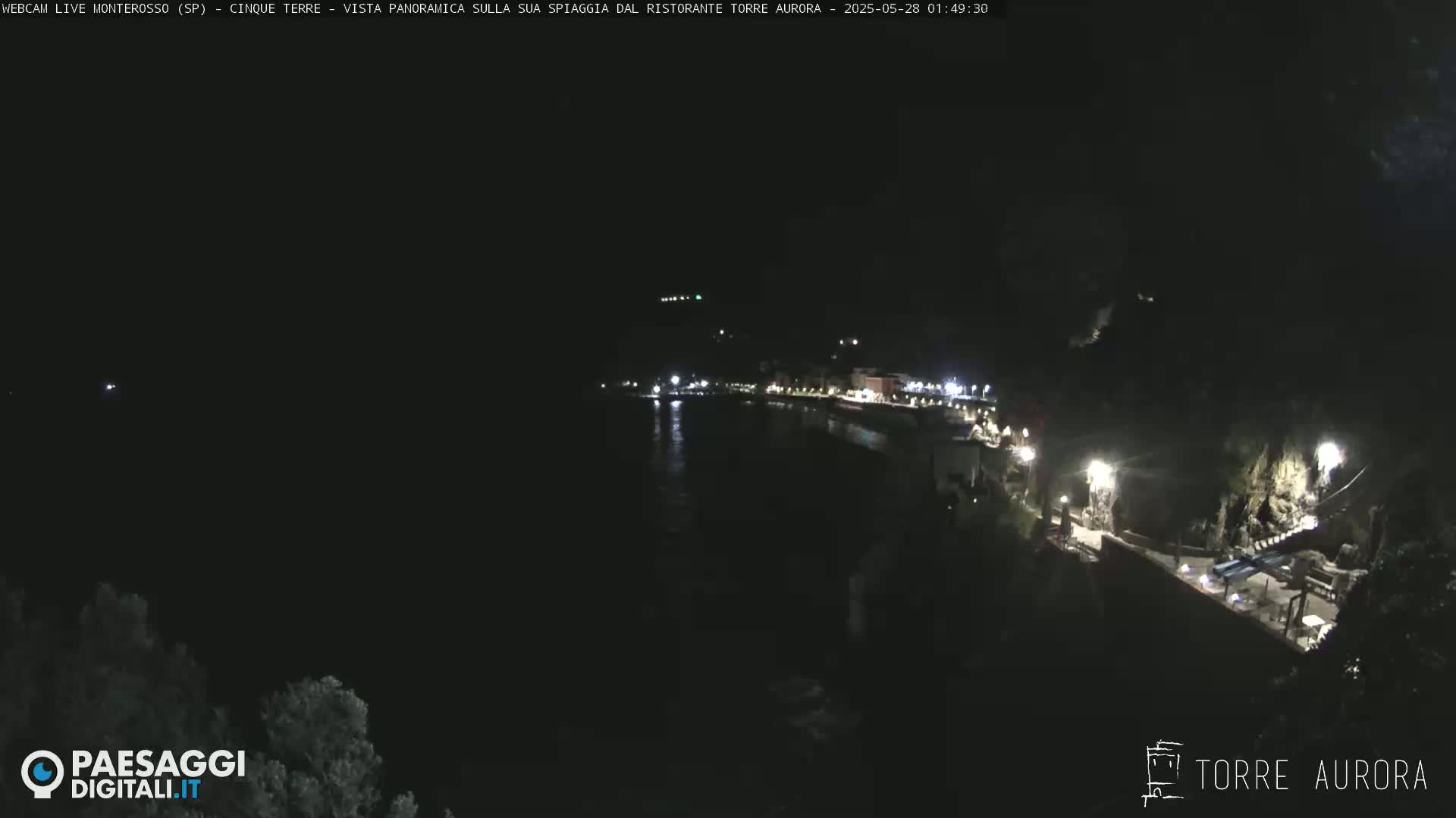 A nighttime, panoramic view of a dark coastline with a few illuminated buildings and a body of dark water.