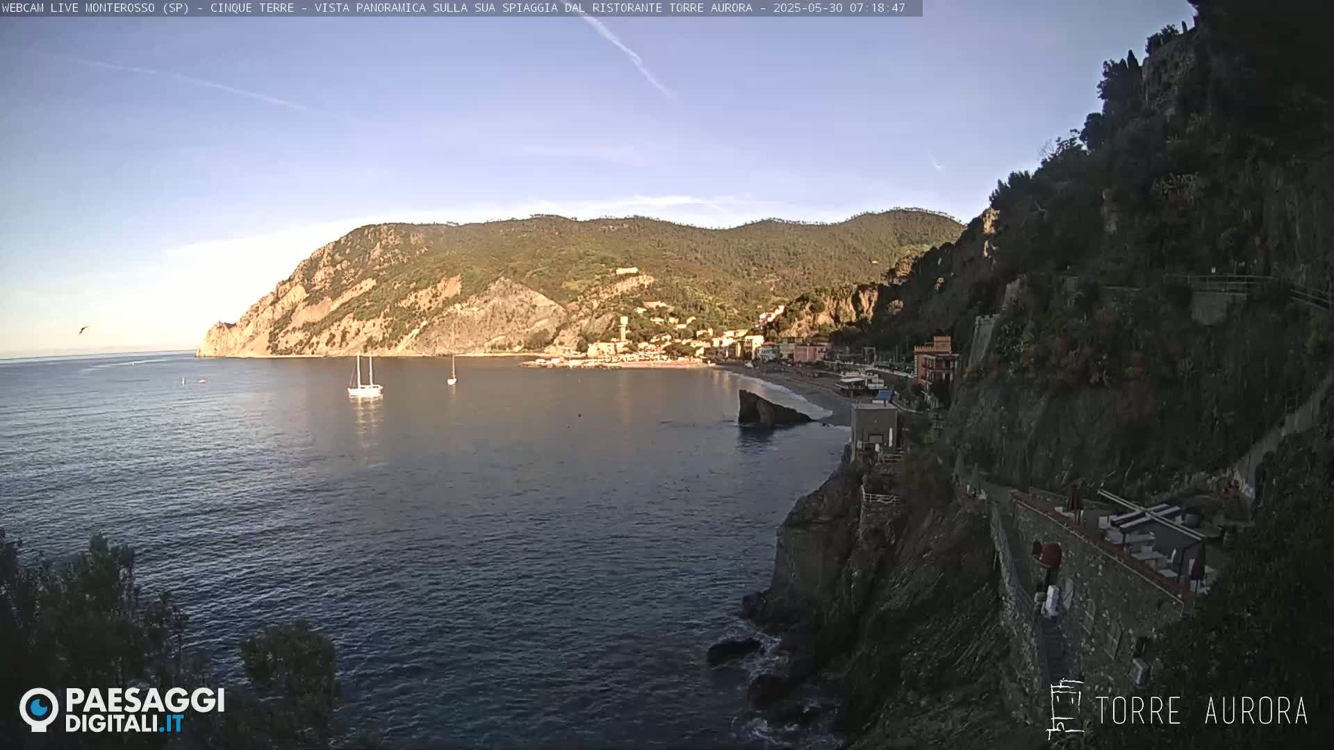 A coastal town nestled between hillsides overlooks a calm sea with a few sailboats under a clear sky.