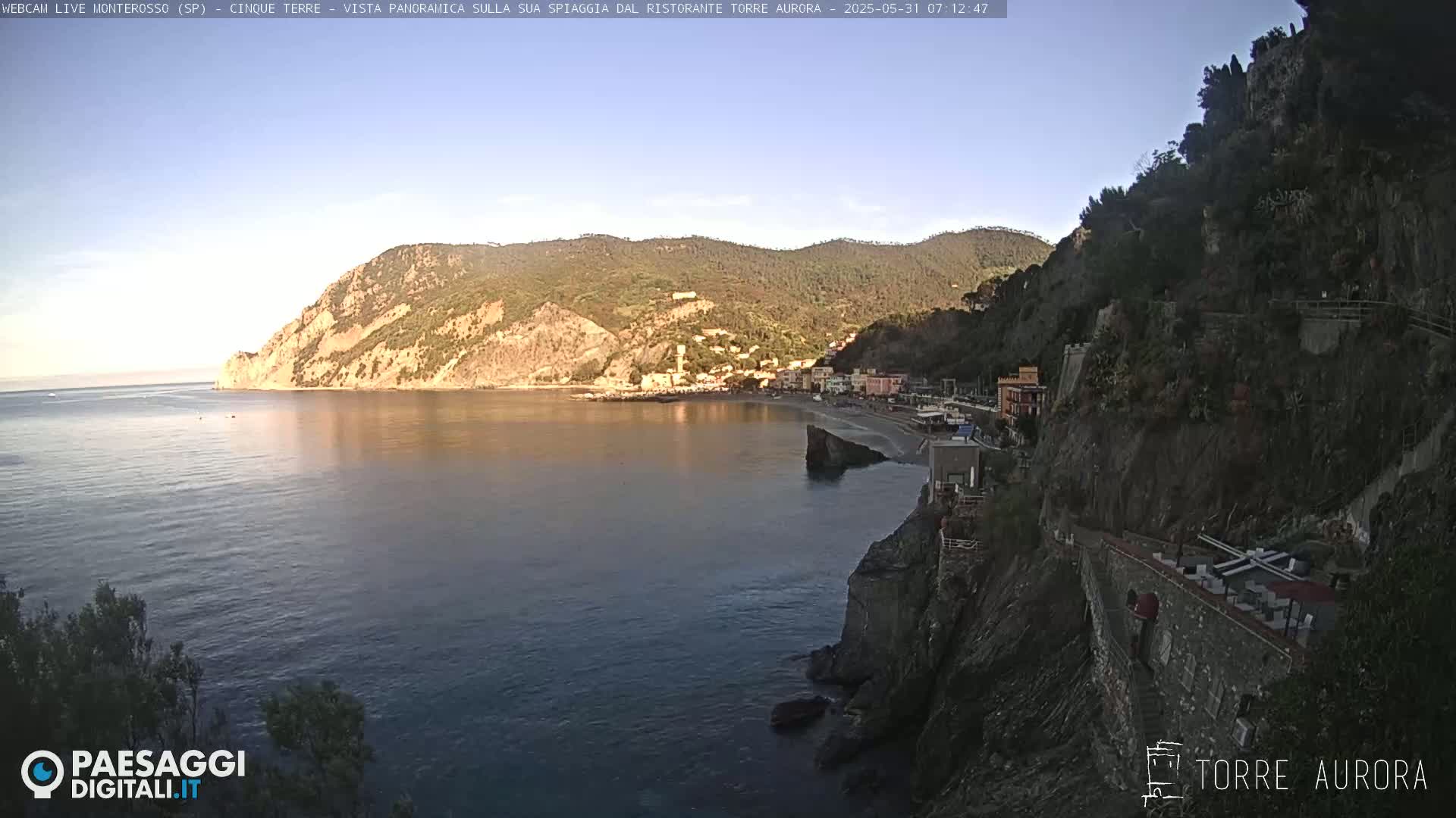 A calm, clear day reveals a coastal town nestled between steep, rocky cliffs, with a small beach and calm waters in front.