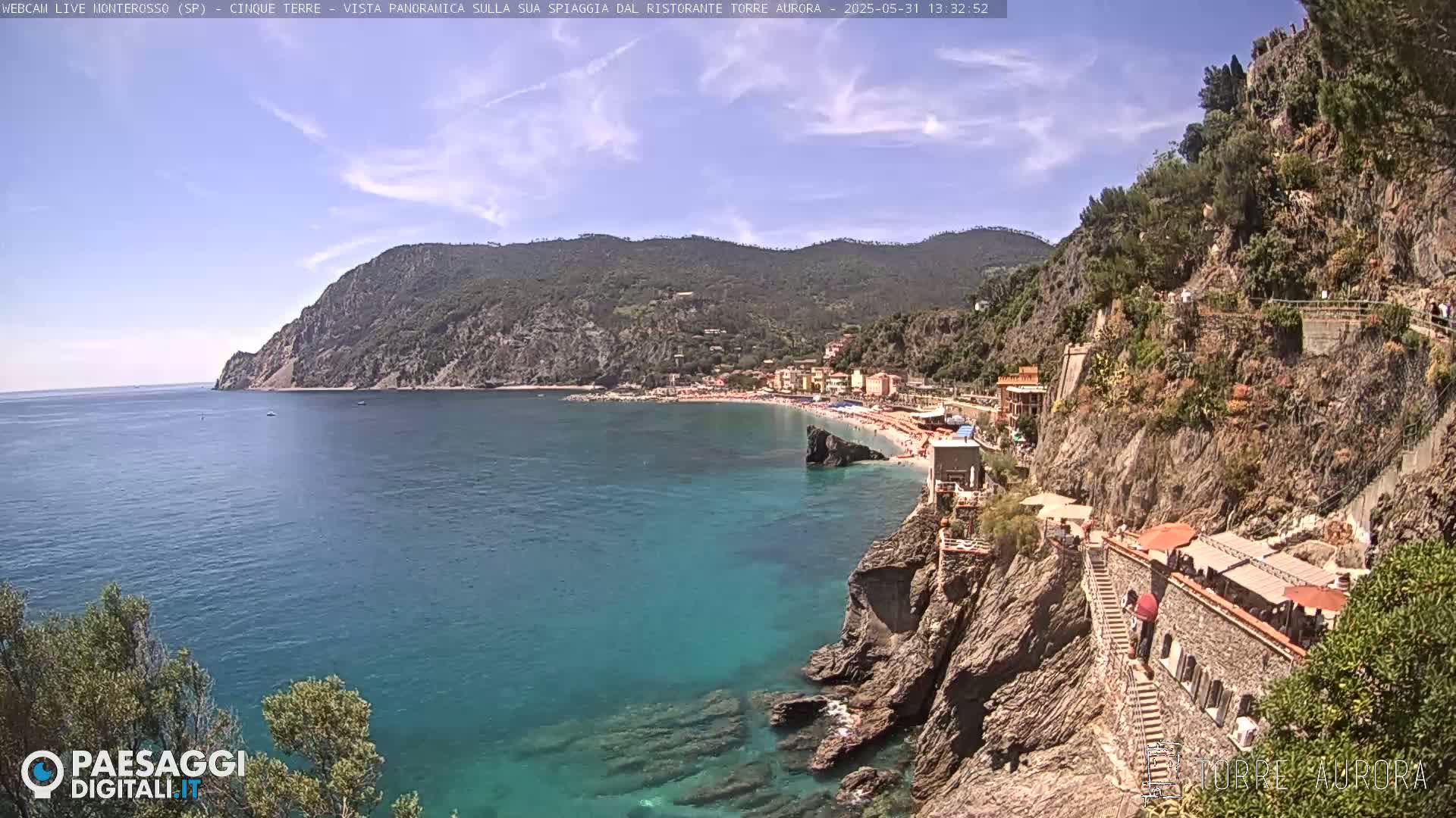 A sunny coastal scene shows a sandy beach and turquoise water nestled between rocky cliffs and hillsides covered in vegetation.