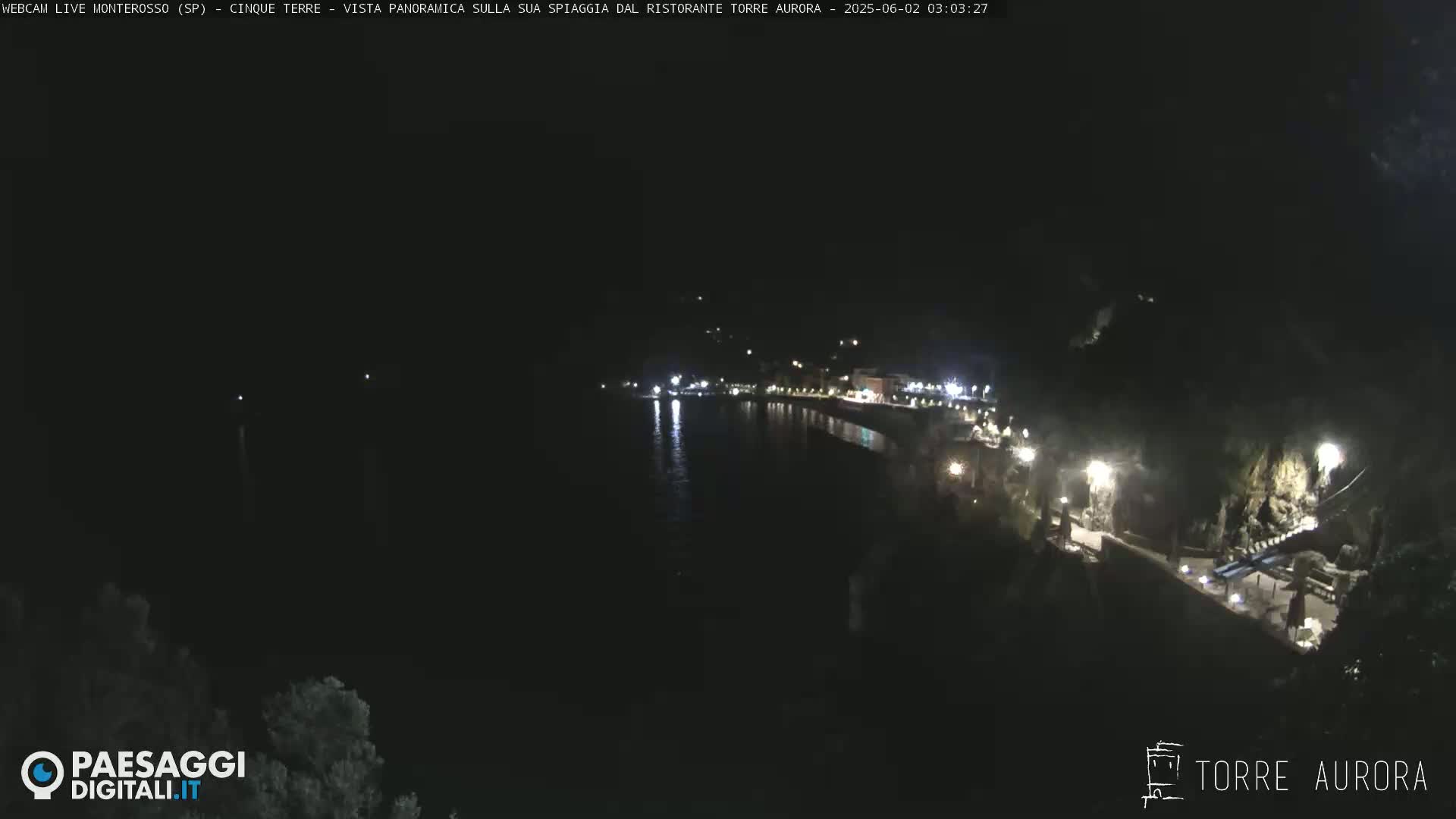 A nighttime, coastal scene shows a dark body of water reflecting the lights of a town nestled on a hillside.
