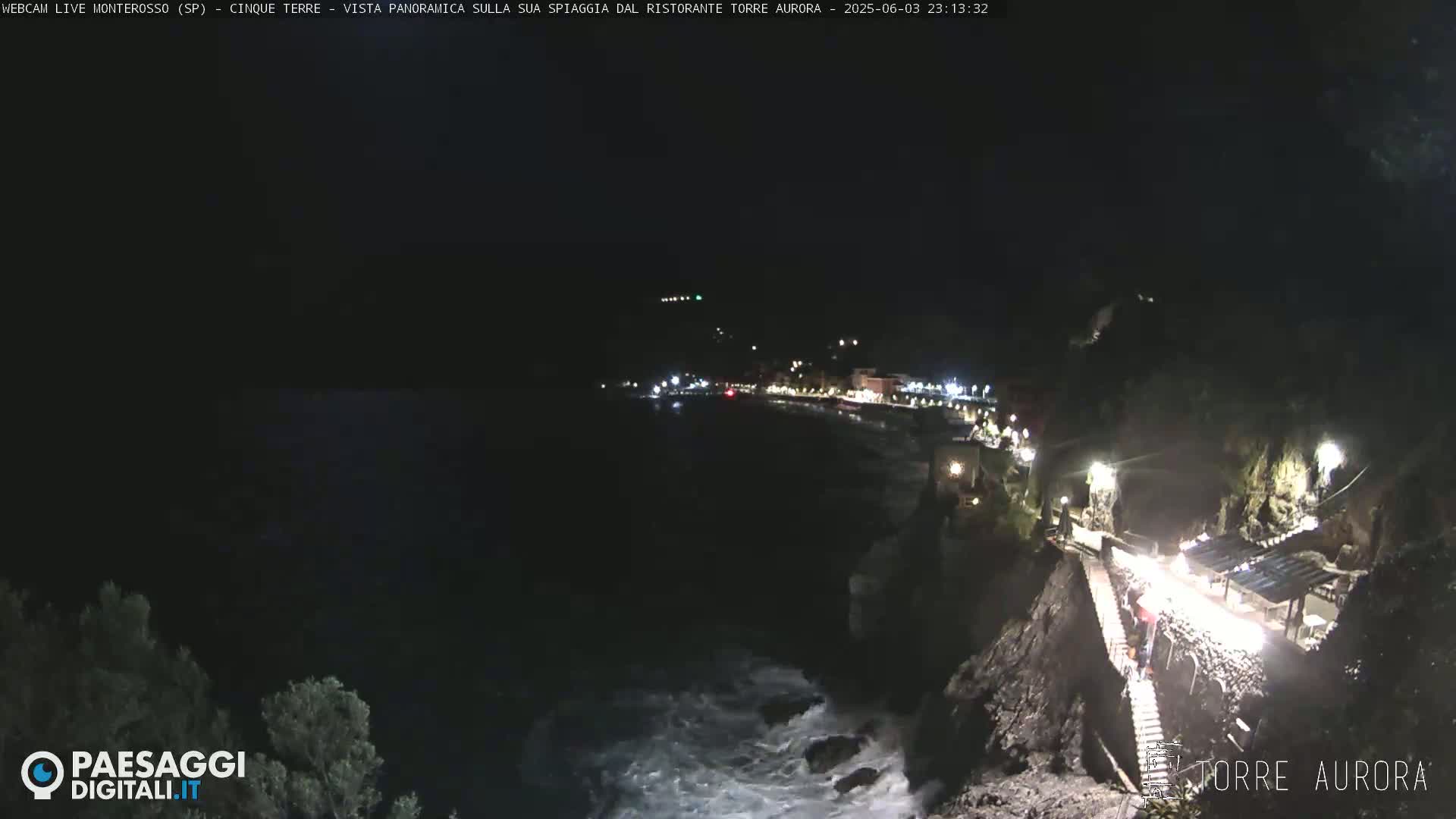 A nighttime coastal scene shows a rocky shoreline with a building lit up, a dark ocean with waves crashing against the rocks, and a town with lights visible in the distance.