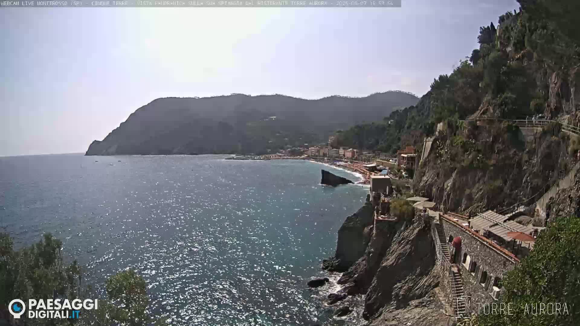 A sunny coastal scene shows a town nestled between steep, rocky cliffs and a calm, sparkling sea.