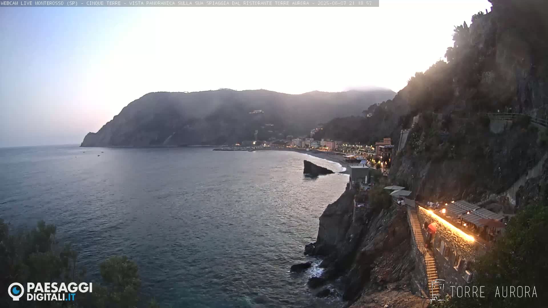 A coastal town at dusk is nestled between a dark cliff and the sea, with a lit pathway leading down to the beach.