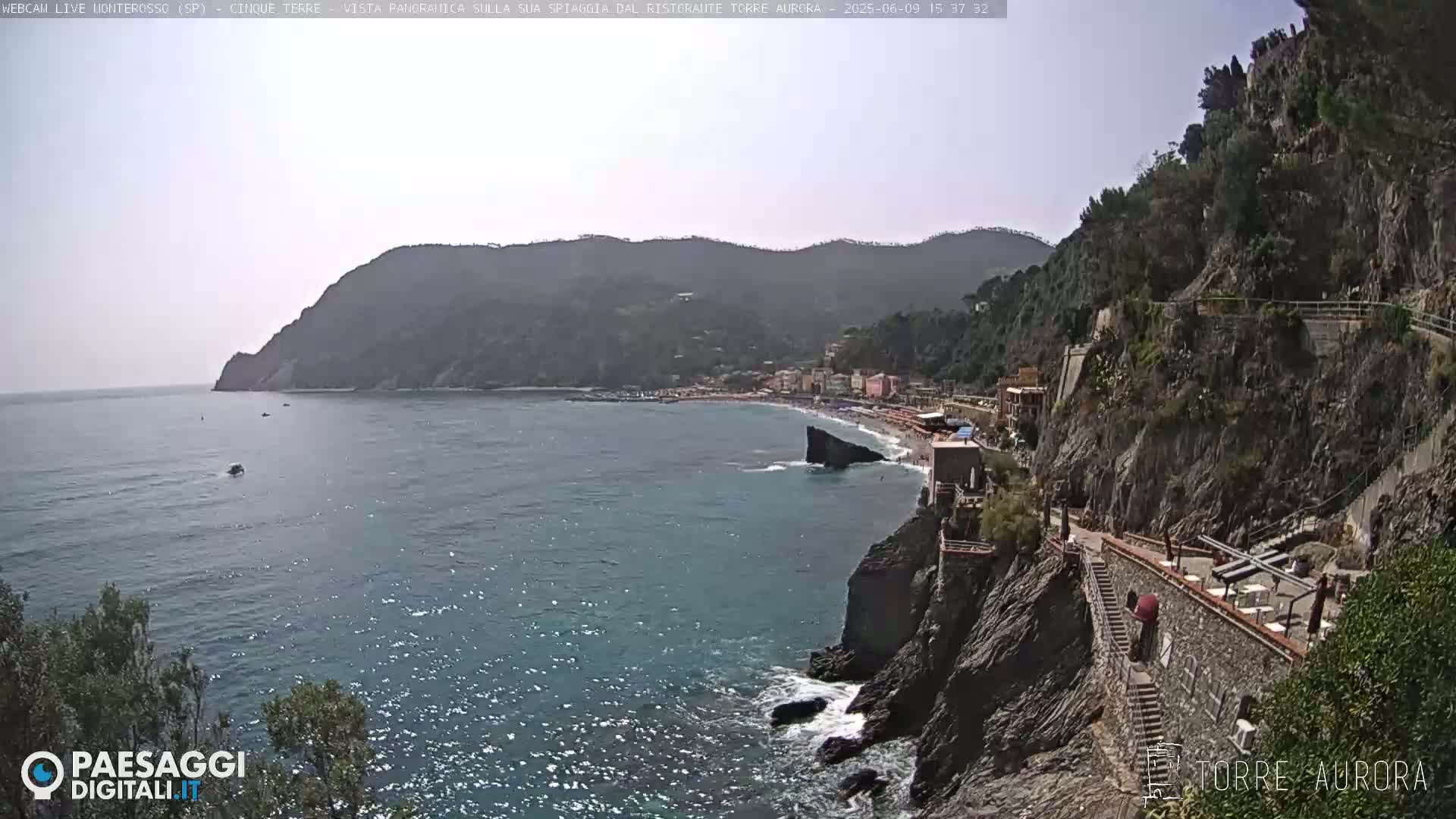 A sunny coastal view shows a rocky cliffside with a pathway and buildings overlooking a calm blue sea and a sandy beach below, backed by distant mountains.