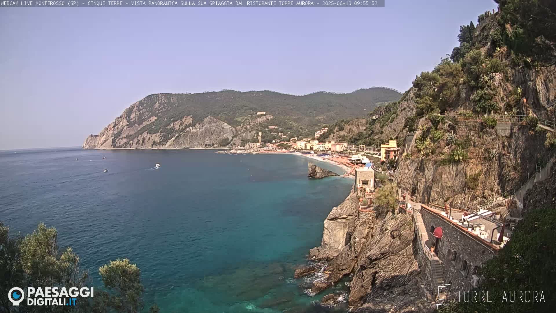 A clear, sunny day reveals a picturesque coastal town nestled between steep cliffs and a turquoise sea, with several small boats visible offshore.