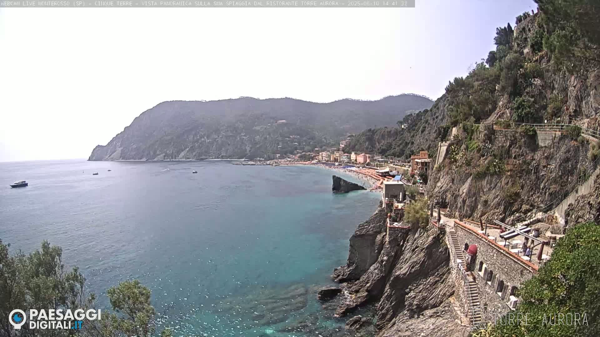A sunny coastal scene shows a turquoise sea, a sandy beach with people, buildings along the shoreline, and a rocky cliff path leading to a seaside restaurant.