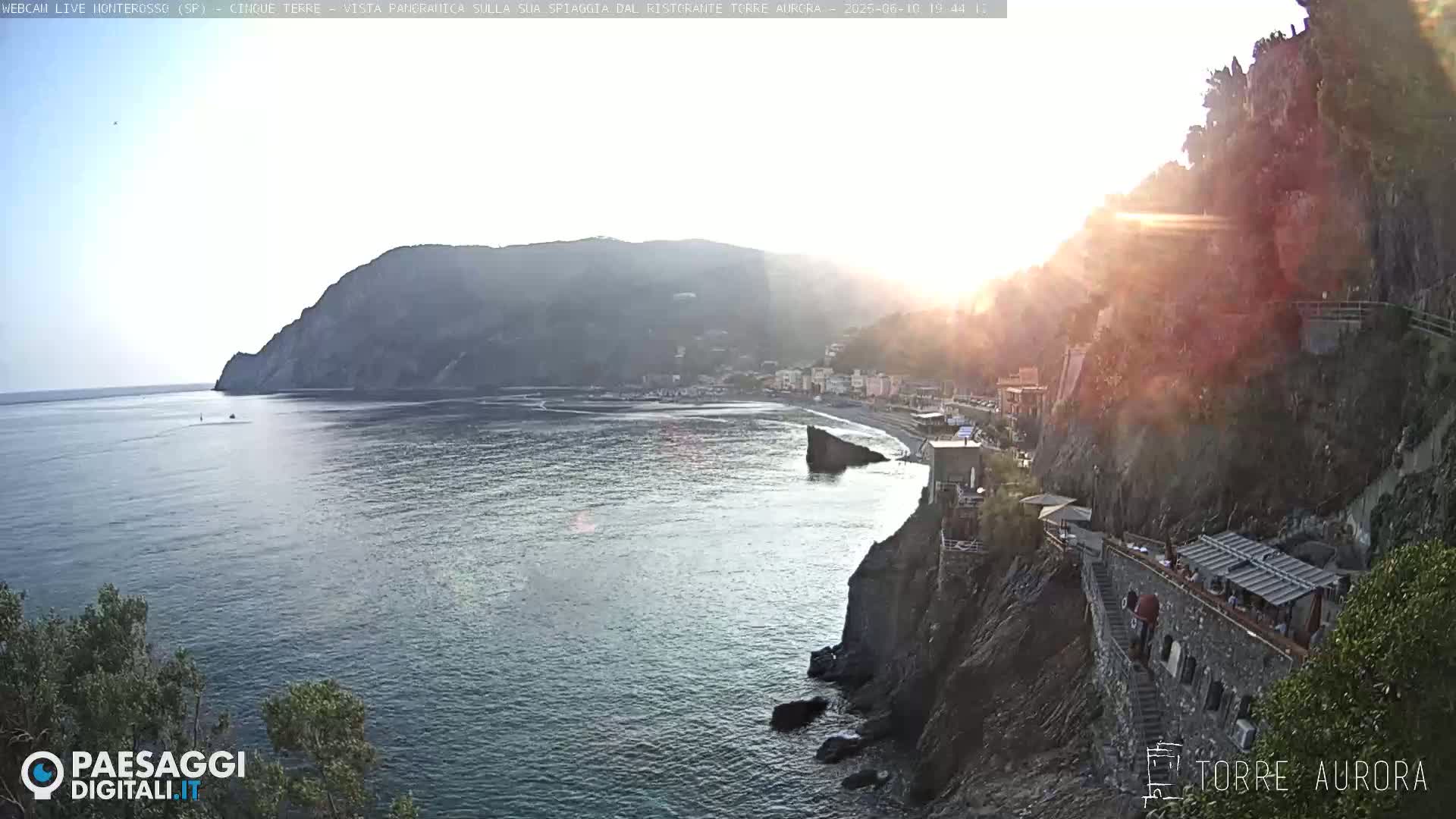 A coastal scene at sunset shows a calm sea, a beach with a small town, and steep cliffs with a building and stairs leading down to the water.
