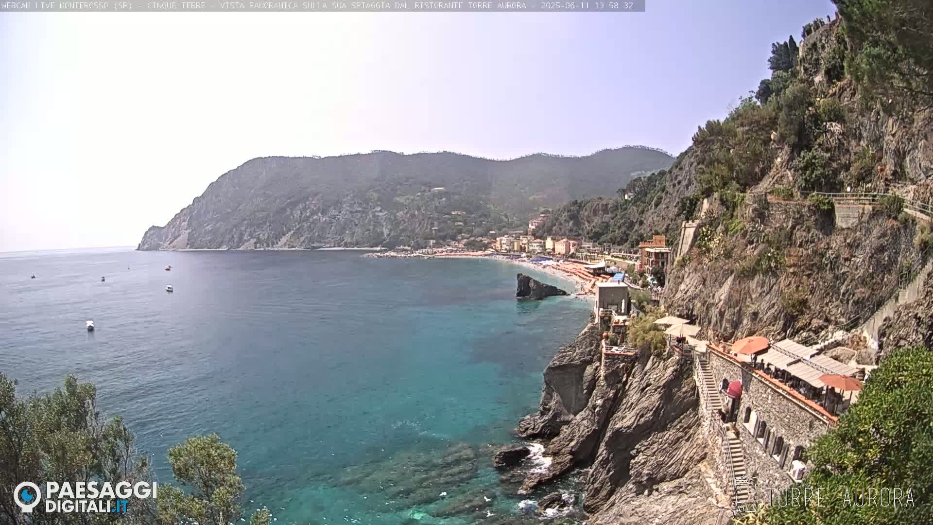 A sunny coastal view shows a beach with people, buildings, and a rocky cliffside path overlooking clear turquoise water.