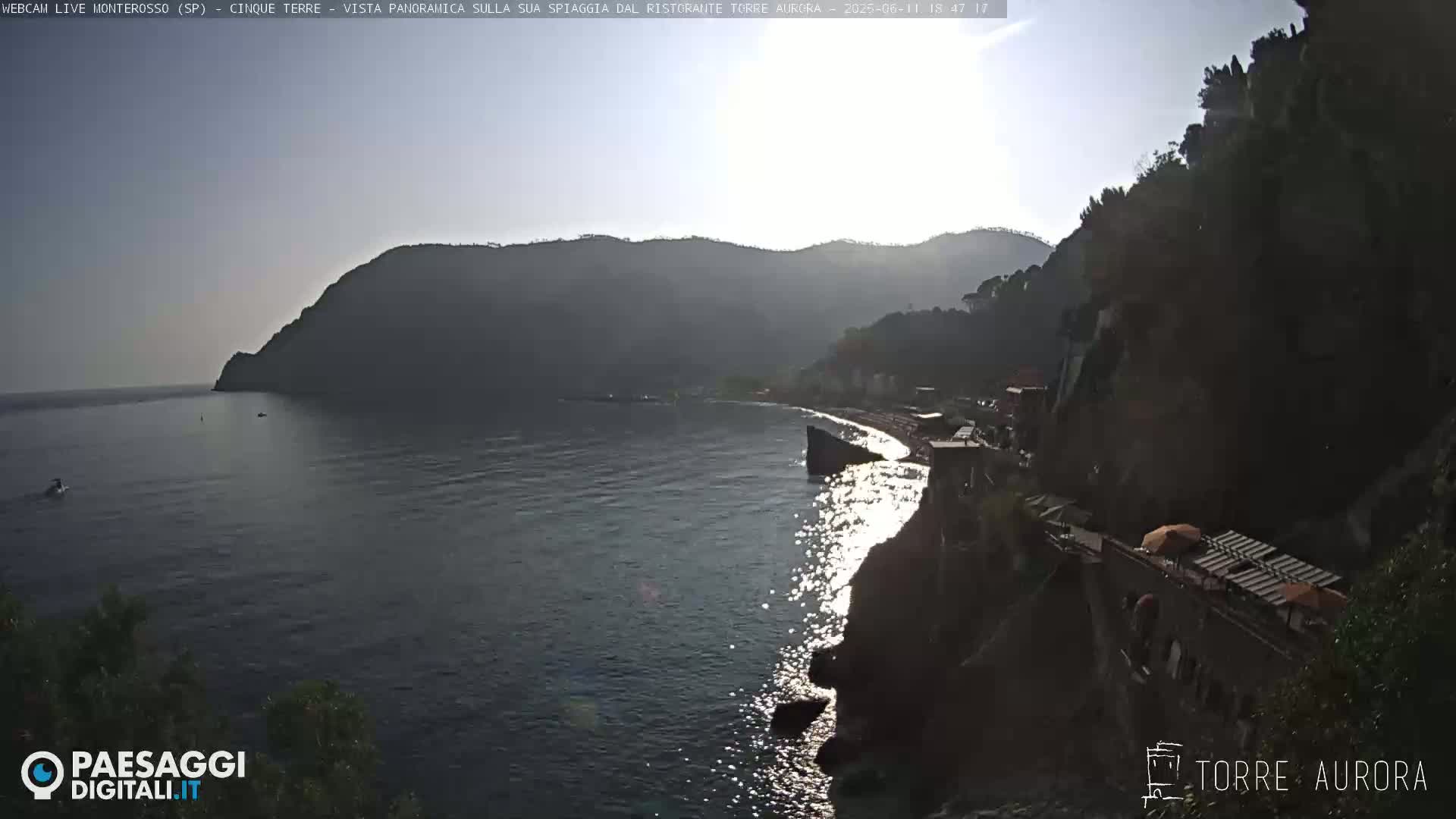 A sunlit coastal scene shows a calm sea reflecting sunlight near a rocky shoreline with buildings and vegetation on the hillside.