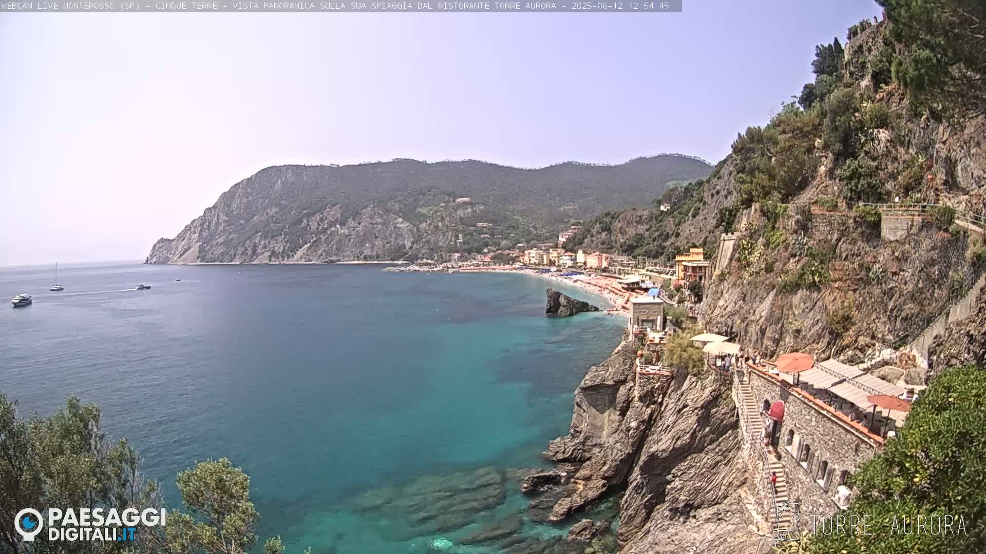 A clear, sunny day overlooks a picturesque coastal town nestled between steep, rocky cliffs and a calm, teal sea.