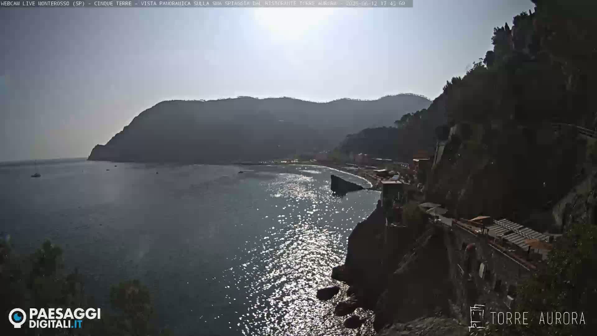 A sunny day reveals a sparkling sea lapping at the rocky shore below a hillside town nestled amongst mountains.