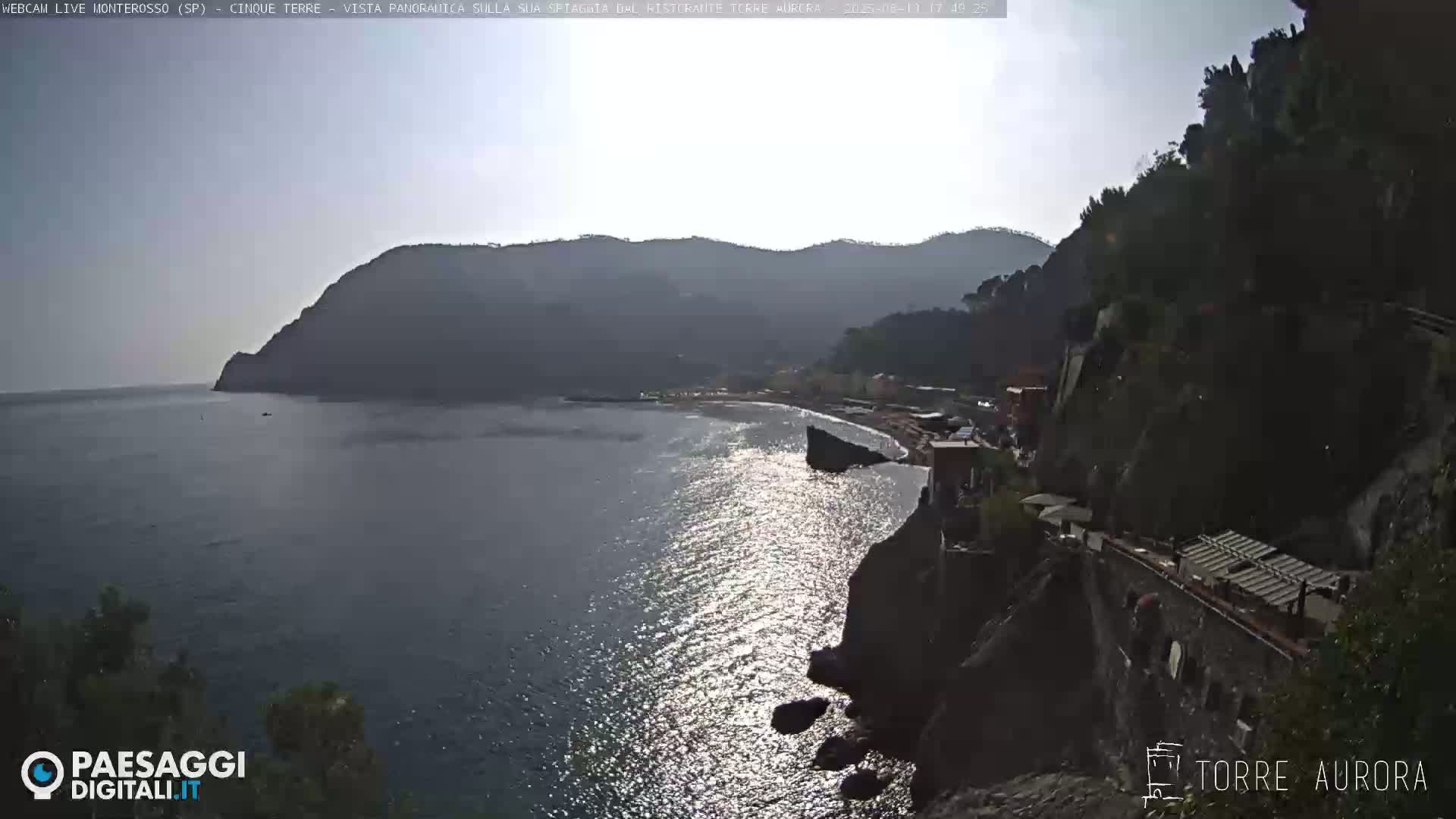 A sunny coastal scene shows a calm sea reflecting sunlight, with a beach and buildings nestled along a rocky hillside.