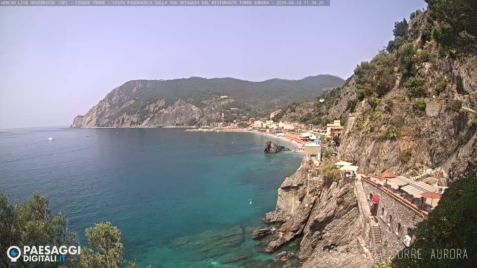 A clear, sunny day overlooks a picturesque coastal town nestled between rocky cliffs and a calm, teal ocean.