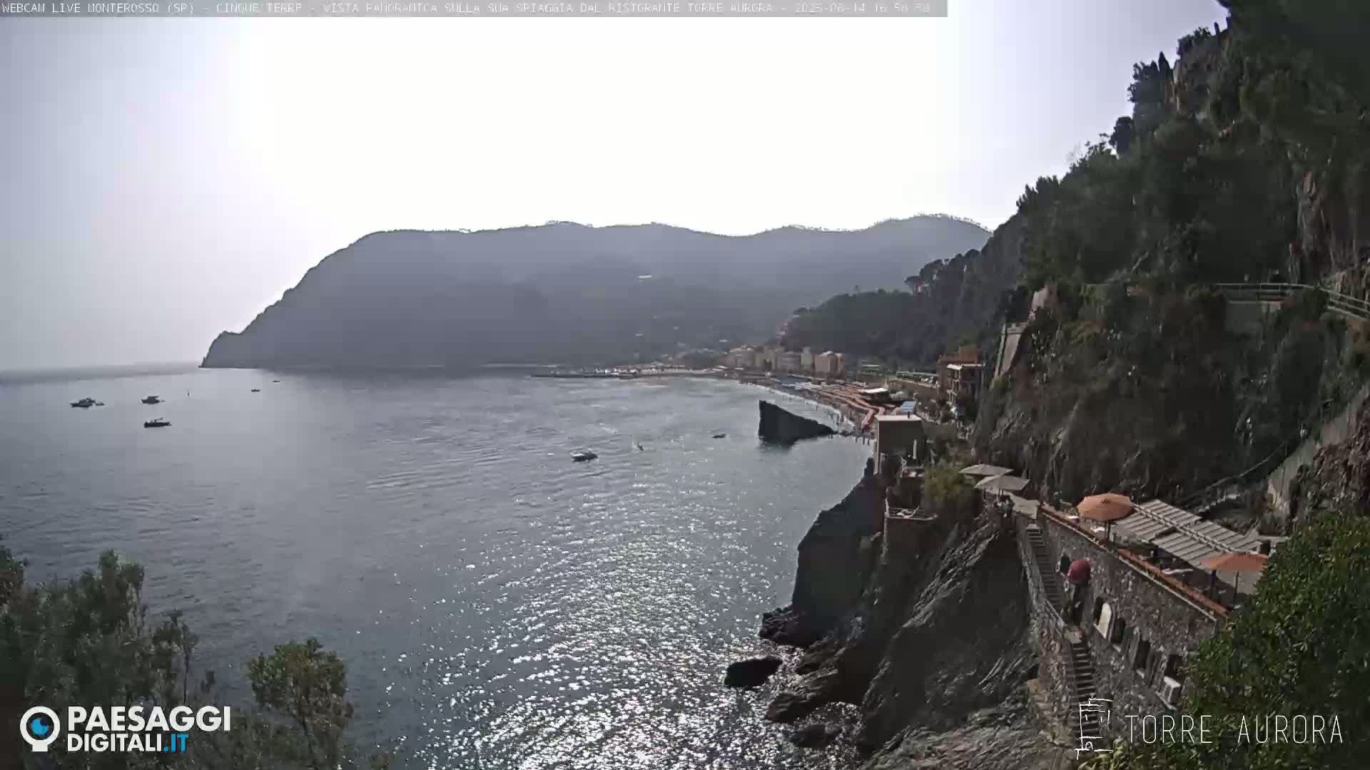A seaside town nestled between hills and a calm sea under a hazy sky is viewed from a high vantage point.
