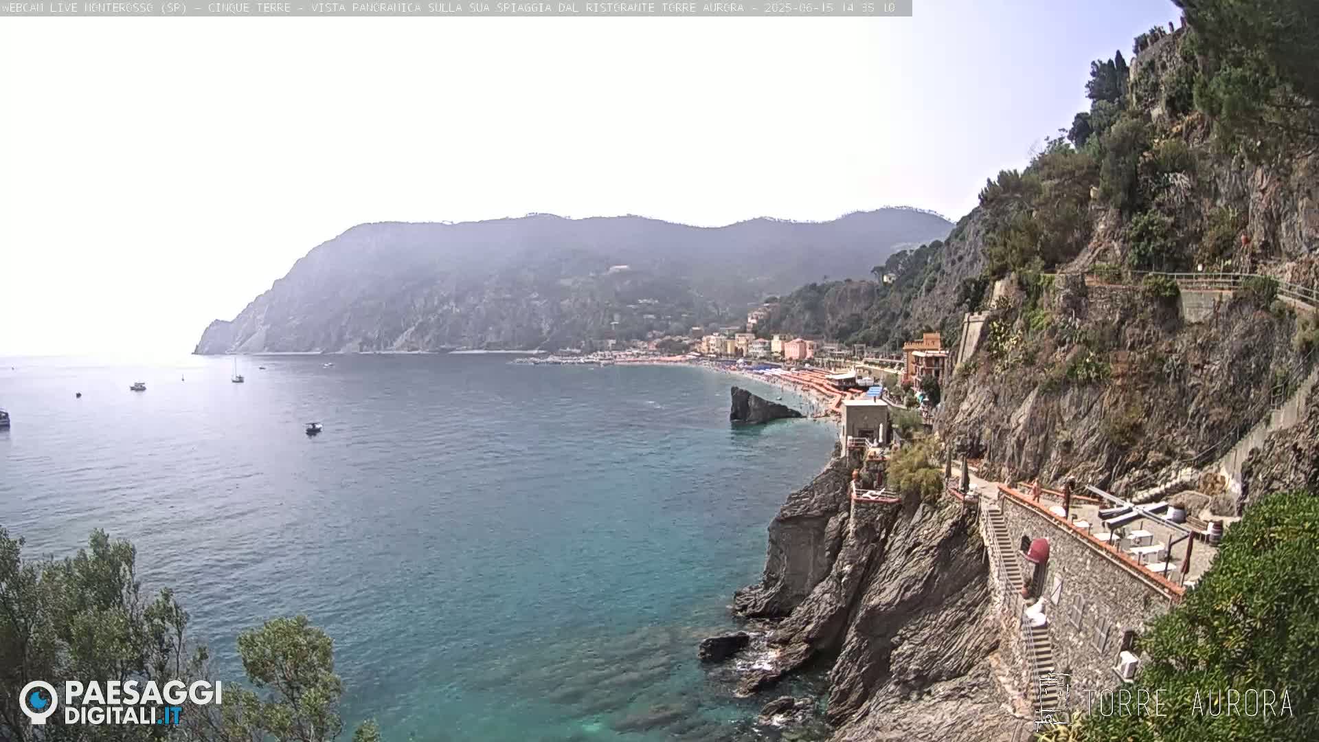 A coastal town with a sandy beach nestled between steep, rocky cliffs overlooks a calm, blue sea on a sunny day.