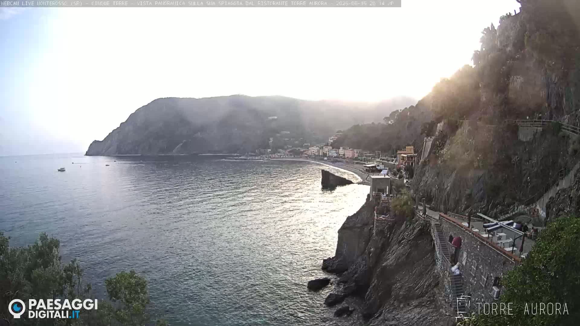 A hazy sunset illuminates a coastal town nestled between steep cliffs overlooking a calm sea.
