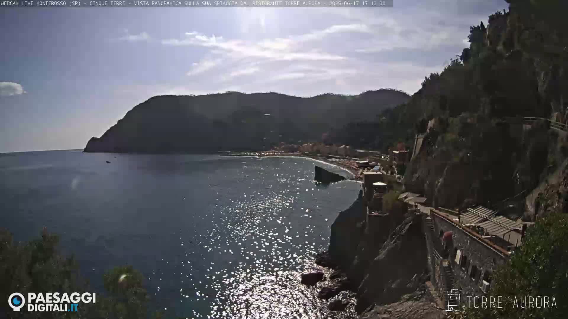 A sunny day reveals a picturesque coastal scene with a small beach nestled between dark, rocky cliffs and a calm, sparkling sea.
