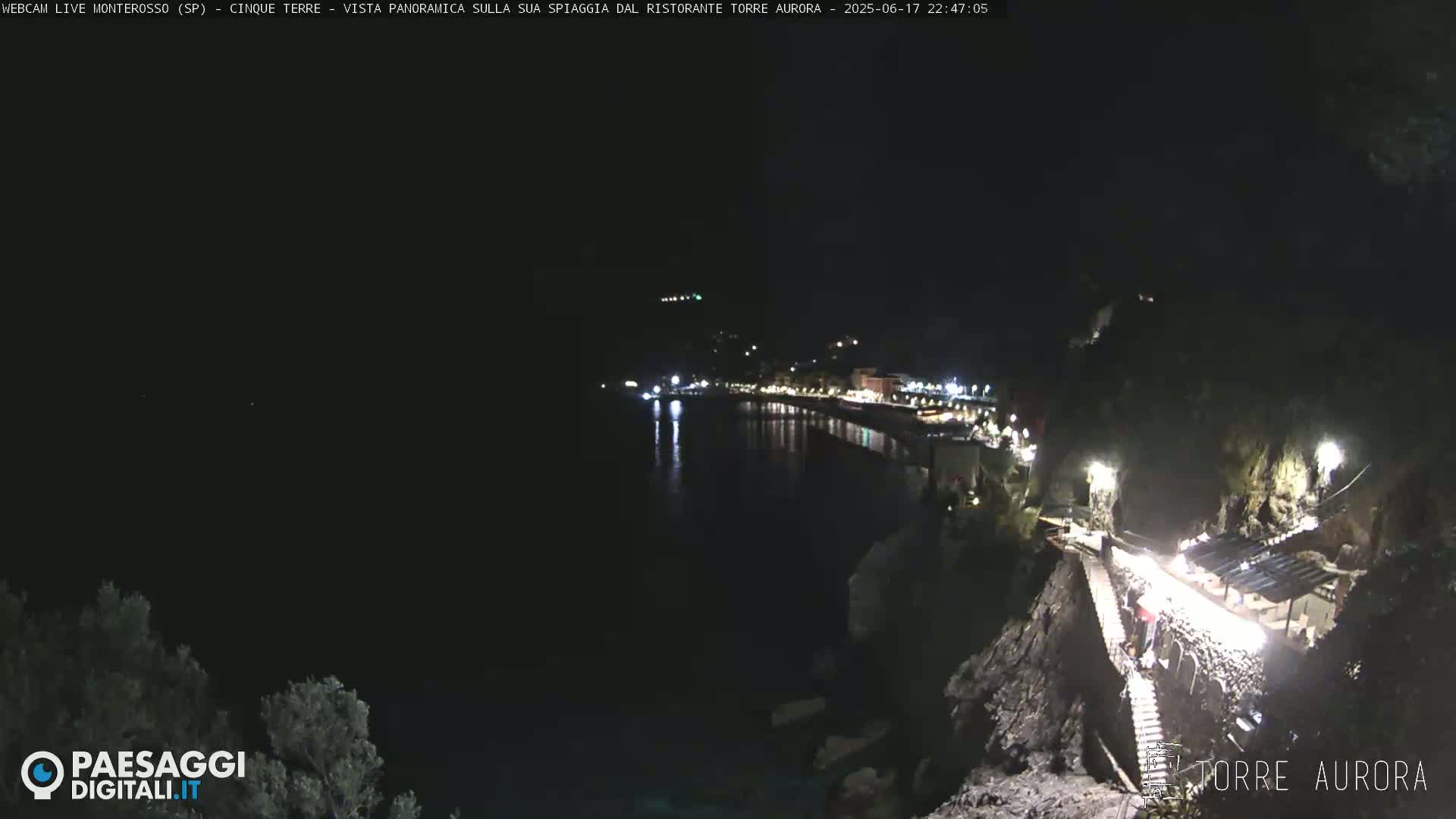 A nighttime, seaside view shows a dark, calm body of water reflecting the lights of a town on the opposite shore, with a lit pathway and structure visible on a rocky cliff in the foreground.