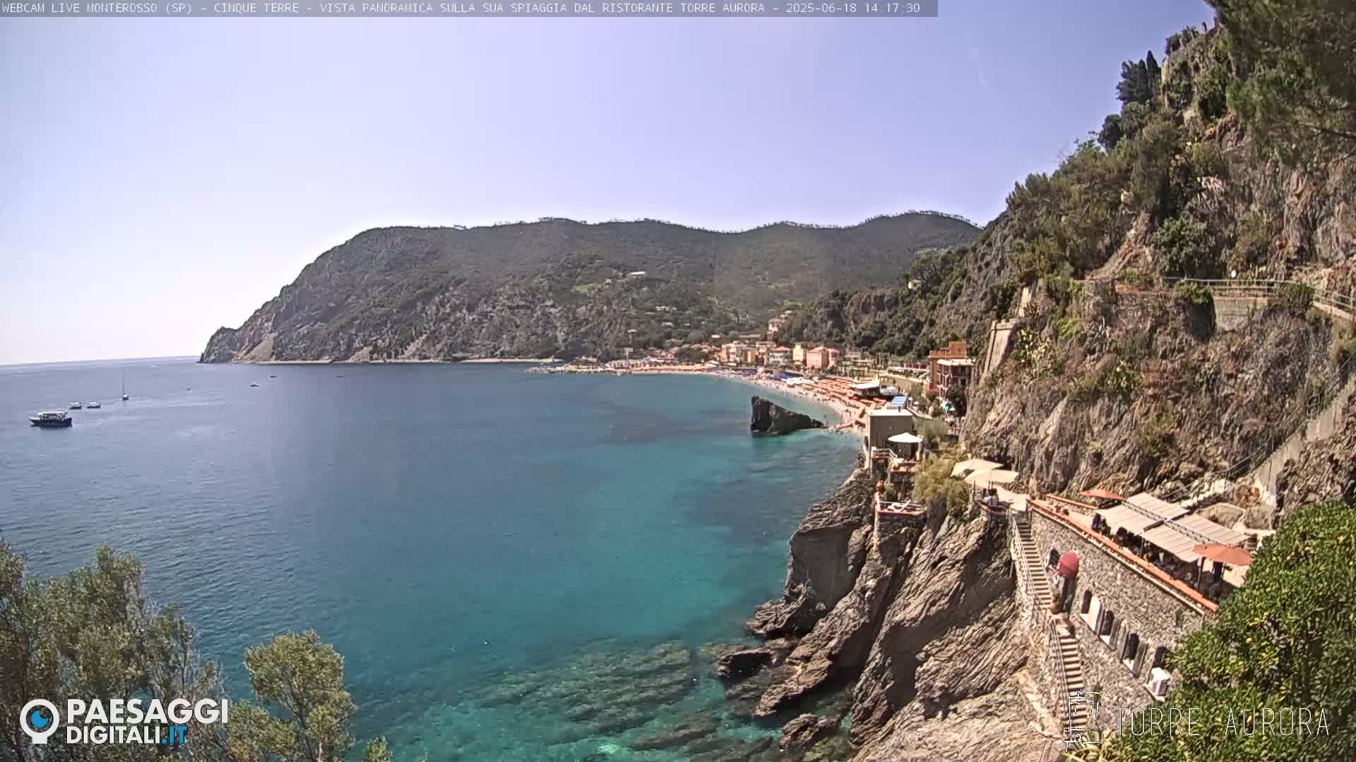 A sunny coastal scene shows a turquoise bay with a sandy beach and buildings nestled against rocky cliffs, under a clear sky.