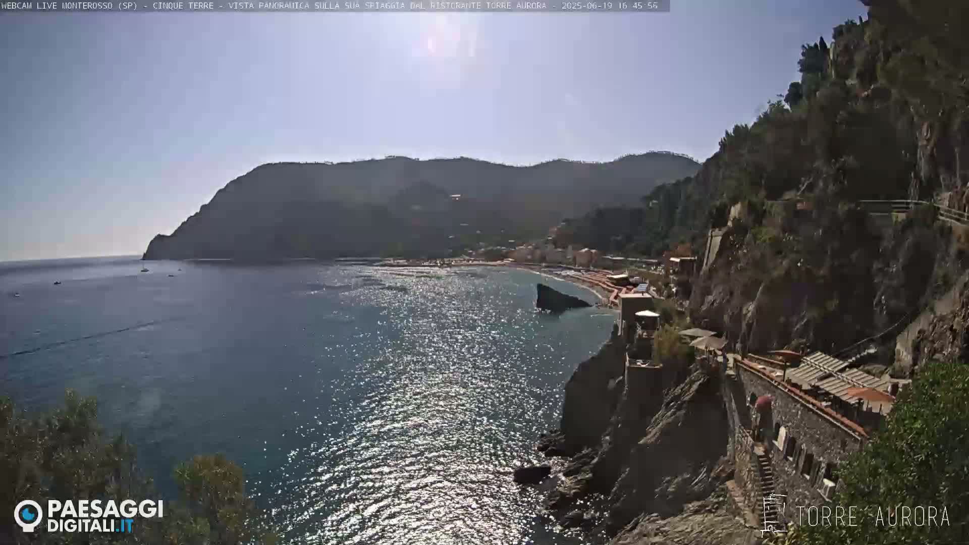 A sunny coastal scene shows a rocky shoreline with a beach and buildings below, overlooking a calm blue sea and mountainous terrain.