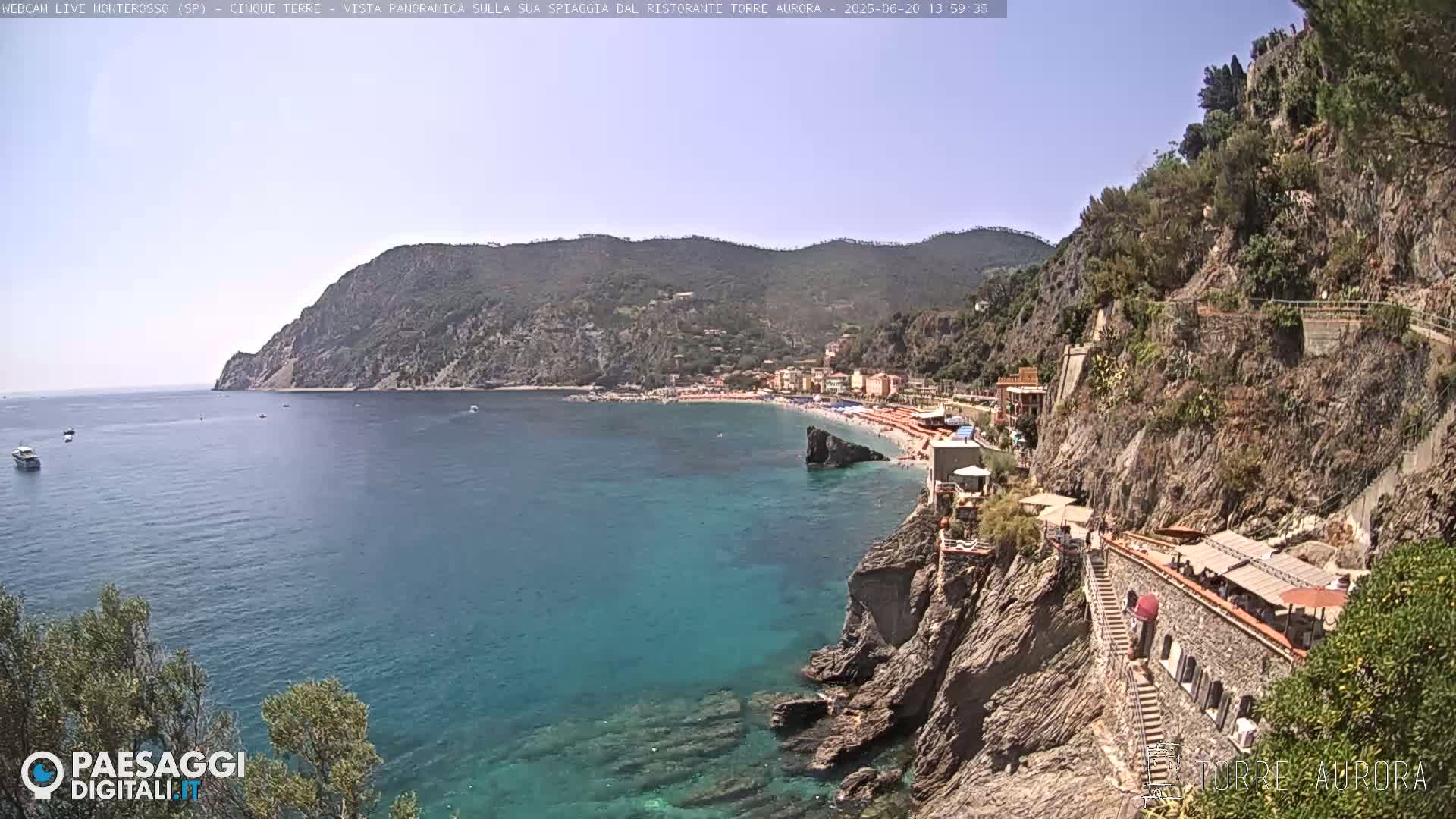 A sunny coastal scene shows a clear blue sea lapping against rocky cliffs, with a beach and buildings nestled in the background.
