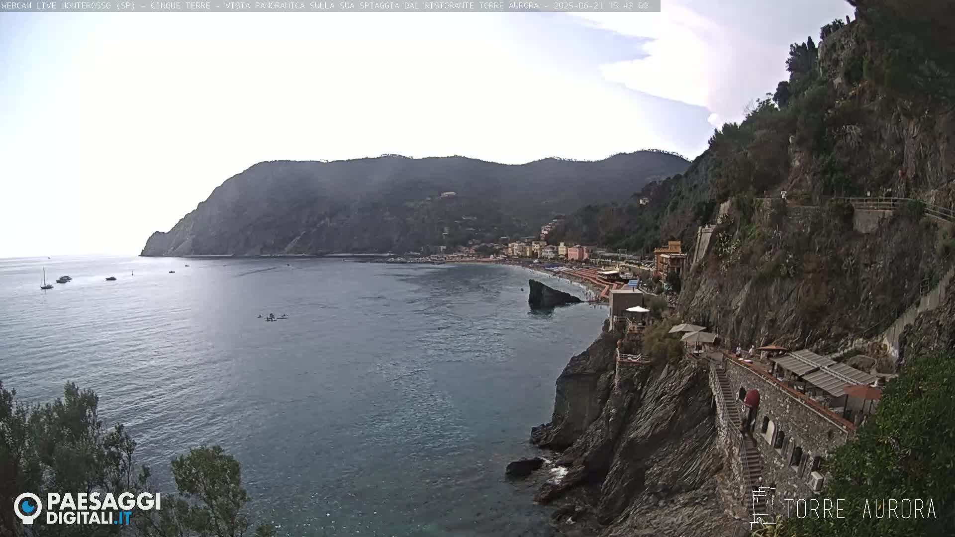 A partly cloudy day shows a coastal town nestled between mountains and a calm sea, with a rocky cliffside path leading to a seaside restaurant.