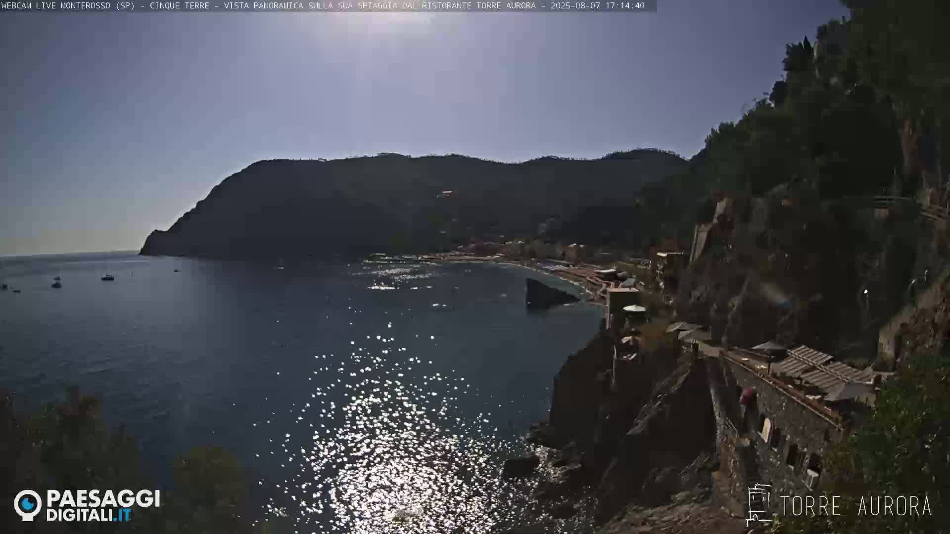 A sunny, high-angle view shows a small, rocky cove with a beach and buildings nestled between steep, dark green hillsides, with calm, sunlit water.