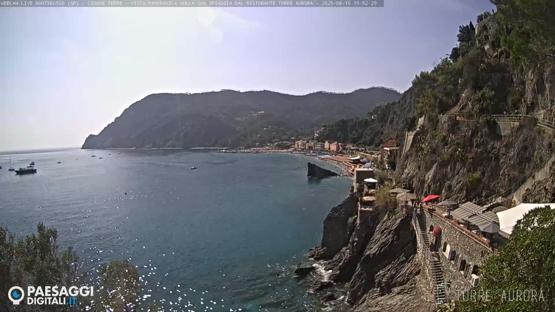 A sunny, high-angle view shows a small, rocky cove with a beach and buildings nestled between steep, dark green hillsides, with calm, sunlit water.