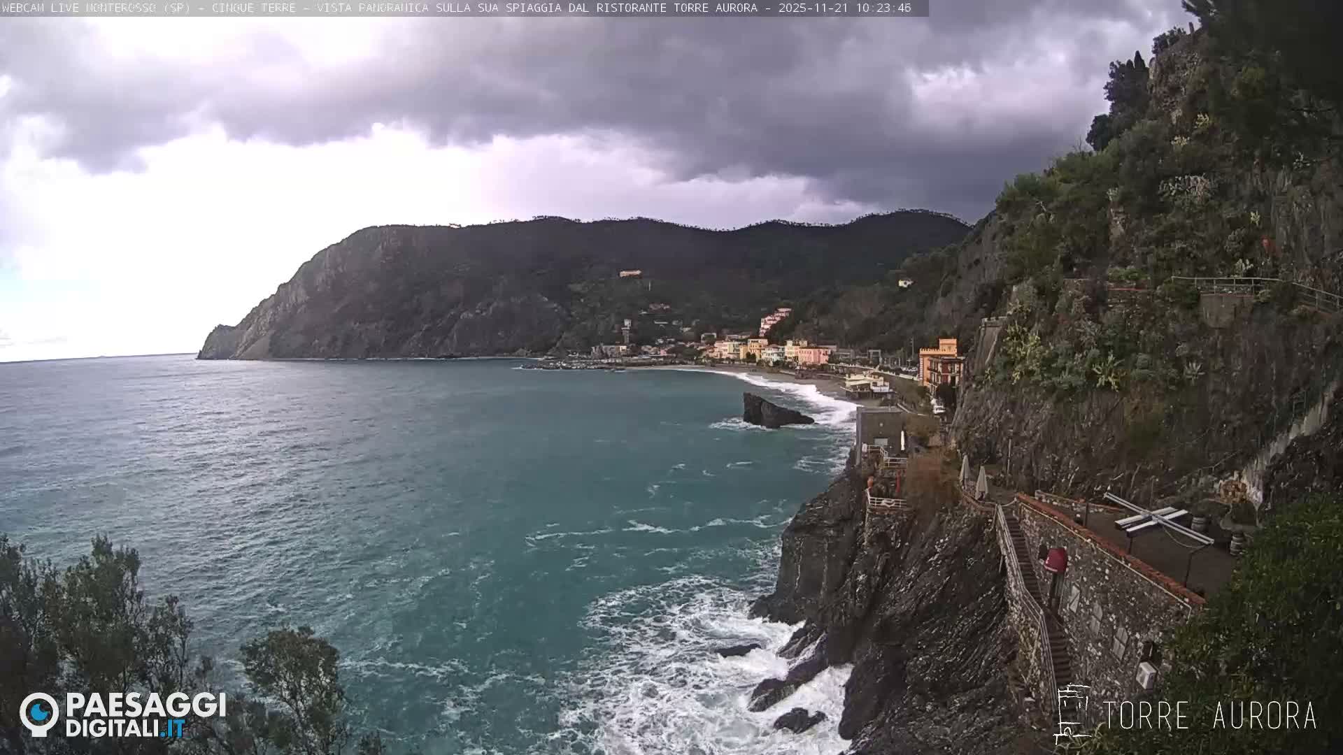 A dramatic coastal landscape under a gloomy, overcast sky shows a colorful Italian village nestled in a cove, with strong waves crashing against rugged cliffs that feature a terraced path in the foreground.