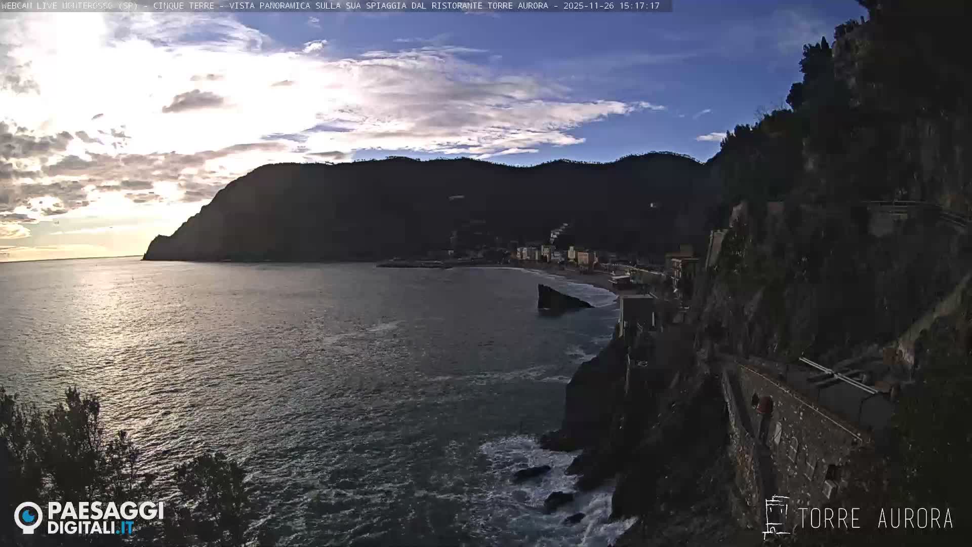 A dramatic coastal landscape under a gloomy, overcast sky shows a colorful Italian village nestled in a cove, with strong waves crashing against rugged cliffs that feature a terraced path in the foreground.