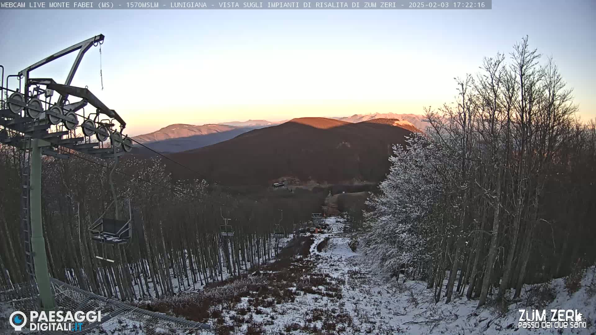 A snow-dusted ski lift ascends a mountain slope at sunset, overlooking a snow-covered forest and distant mountains.