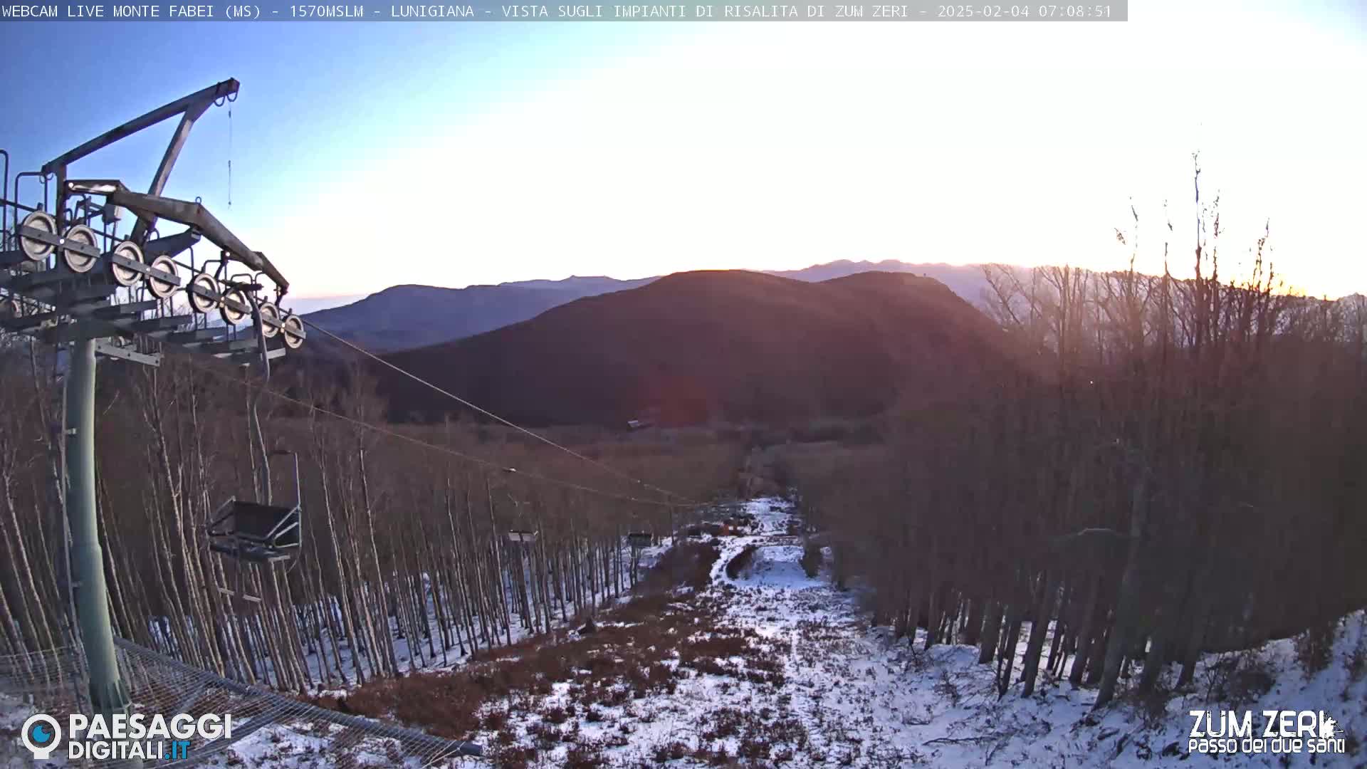 A chairlift ascends a snowy mountain slope at sunrise, passing through a sparsely wooded area with some bare trees.