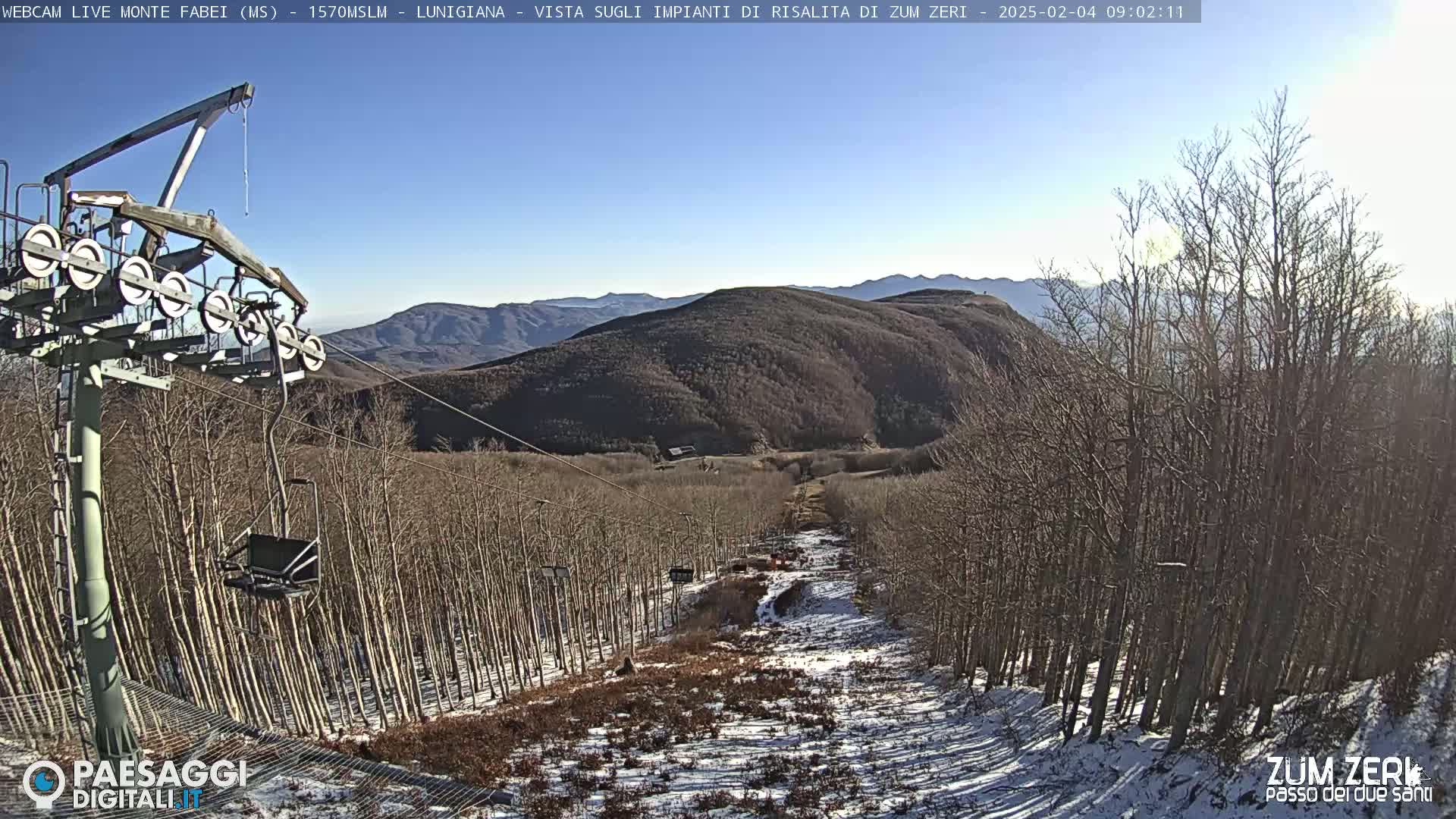 A chairlift ascends a snow-dusted mountainside lined with bare trees under a clear, sunny sky.