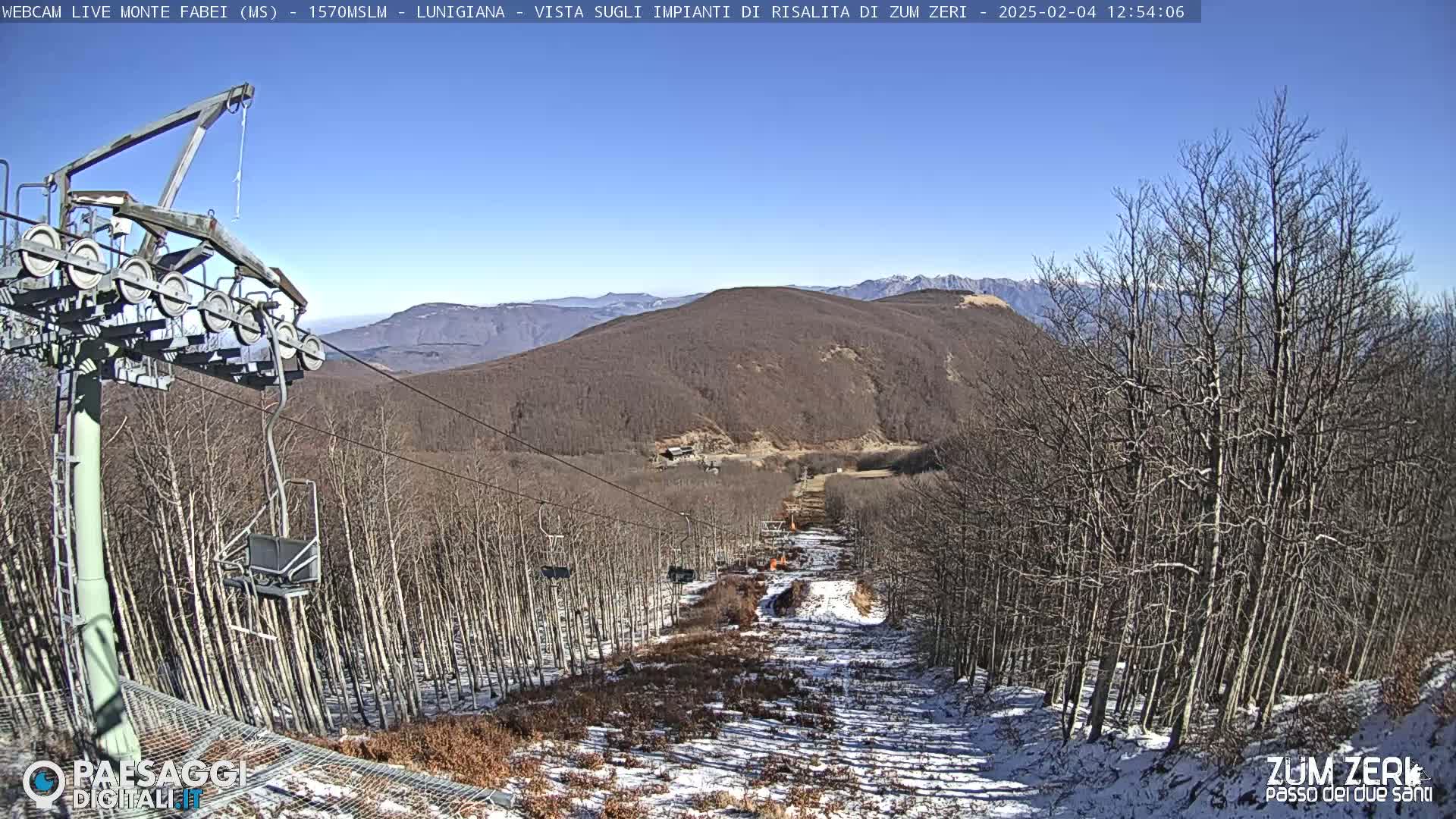 A ski lift ascends a snow-dusted mountainside under a clear blue sky, passing through a forest of leafless trees.
