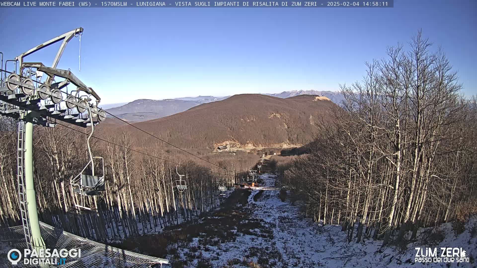 A chairlift ascends a snow-dusted mountainside through a forest of bare trees under a clear blue sky.
