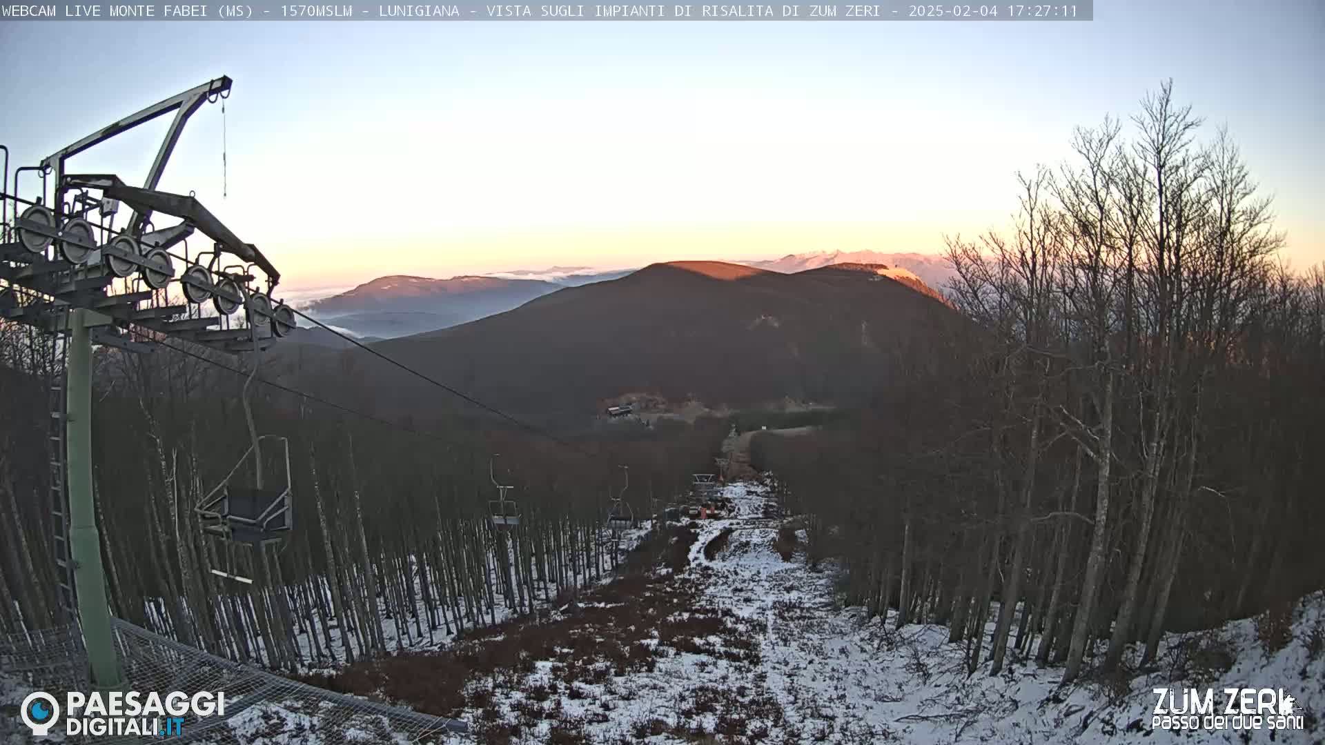 A chairlift ascends a snowy mountain slope at sunset, with a valley and distant mountains visible in the background.