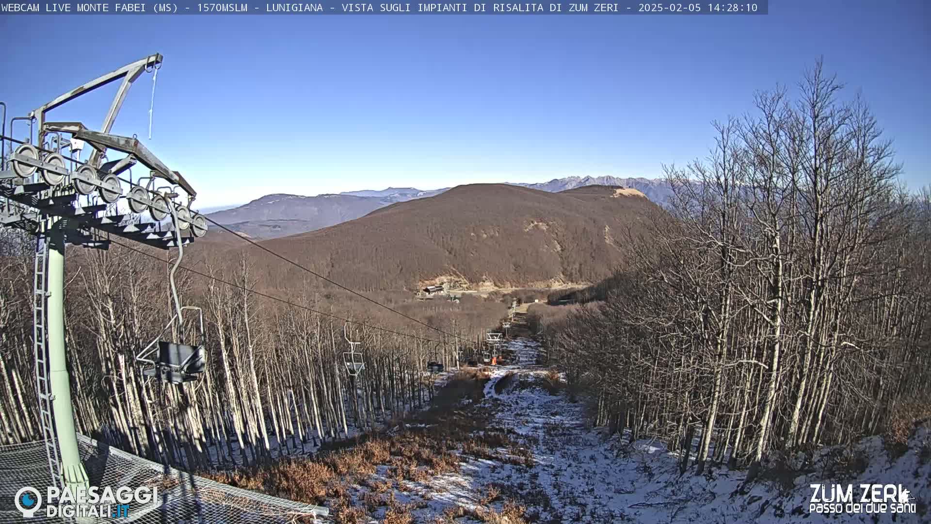 A chairlift and ski slope are visible, with a mostly barren, snow-dusted forest and distant mountains under a clear blue sky.