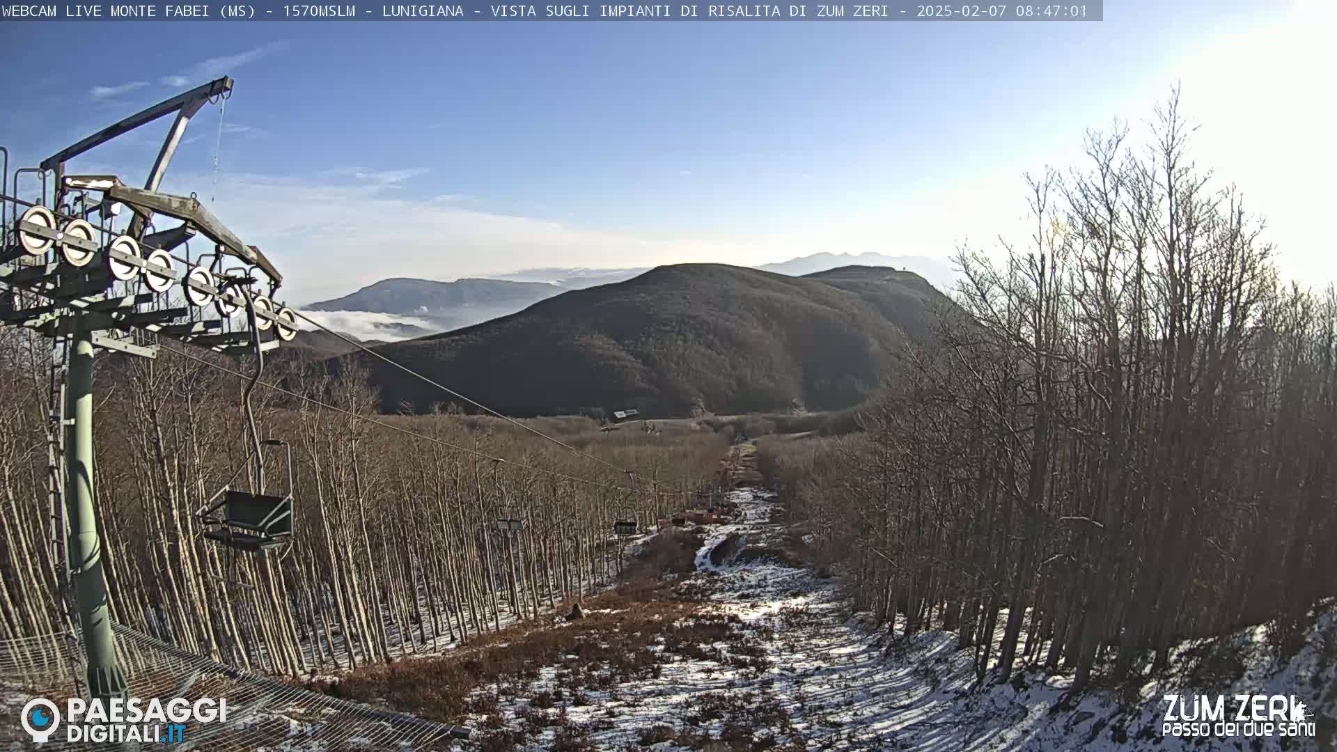 A chairlift ascends a snow-dusted mountainside lined with bare trees under a clear, sunny sky.