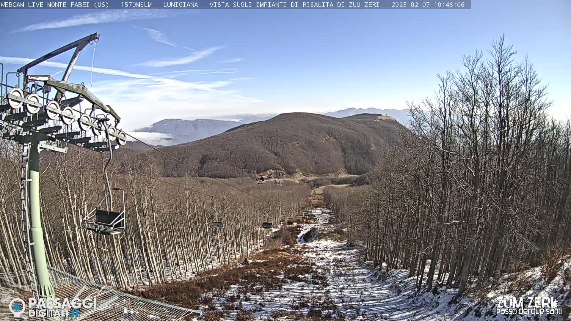 A ski lift chair ascends a mountain slope covered in snow and bare, leafless trees under a clear blue sky.
