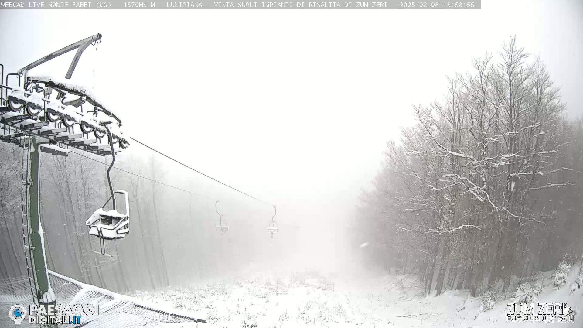 A snow-covered ski lift chair hangs from a cable, with snow falling in a hazy, snowy landscape of snow-covered trees.