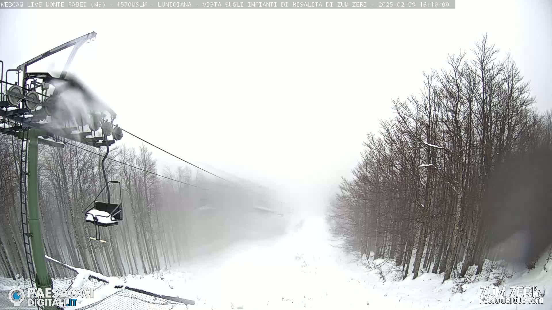 A snow-covered ski lift operates in heavy snow, with bare trees lining a snowy slope.